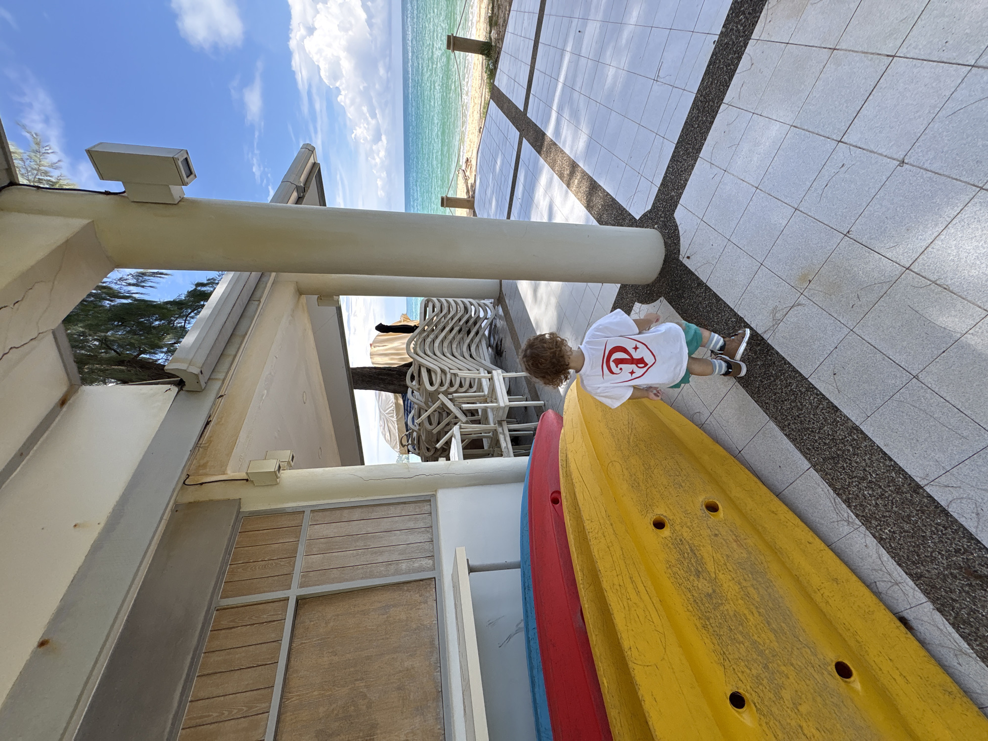 A young child wearing a white t-shirt with a red emblem and green shorts stands on a tiled patio facing the ocean. The patio has a dark gray grout between the light gray tiles. To the left of the child are three kayaks, yellow, red, and blue, stacked from bottom to top. The child is looking out towards the turquoise ocean and a sandy beach. In the background, there is a covered structure with white pillars and a white roof, and stacks of white chairs. The sky is blue with scattered white clouds. The image appears to have been taken in Choeng Thale, Thailand, given the tropical setting and the presence of kayaks often rented at beach resorts.