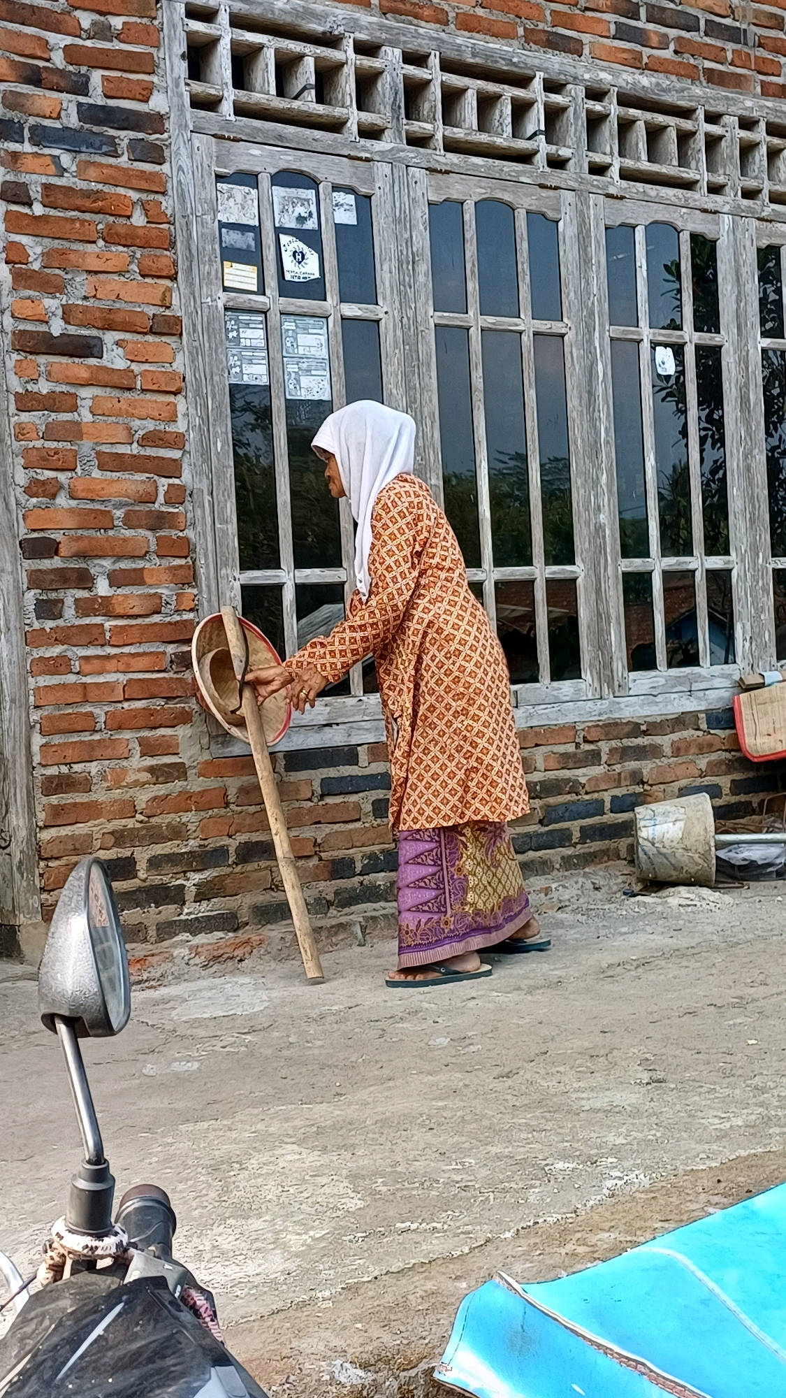 In the city of Pringsurat, Indonesia, a woman wearing a white hijab and a patterned orange and brown jacket over a purple sarong is standing outdoors next to a brick building. She is holding a large, round, woven tool with a wooden handle, possibly for winnowing grain, and appears to be in the process of using it. The building has a large wooden-framed window with multiple panes. The ground is a concrete surface. In the foreground, a motorcycle mirror and handlebars are visible, and to the right, a portion of a bright blue object, possibly a boat or container, is present. The scene suggests a rural or semi-rural setting.