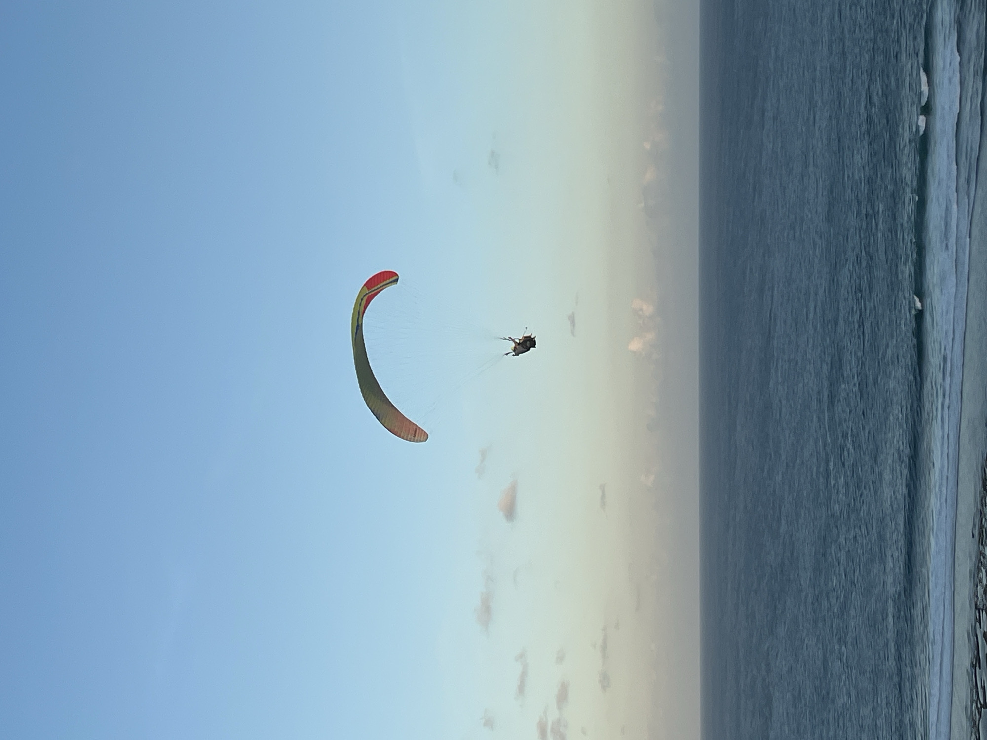 An individual paraglides over the ocean off the coast of Kuta Selatan, Indonesia, under a clear blue sky. The paraglider, with a colorful canopy featuring red, yellow, and green sections, is suspended in the air, connected by lines to the pilot. The vast expanse of the ocean stretches out below, with gentle waves breaking near the shore. The horizon line is visible, separating the sea from the sky, with a few scattered clouds near the lower portion of the atmosphere. The scene appears to be captured during daylight hours.