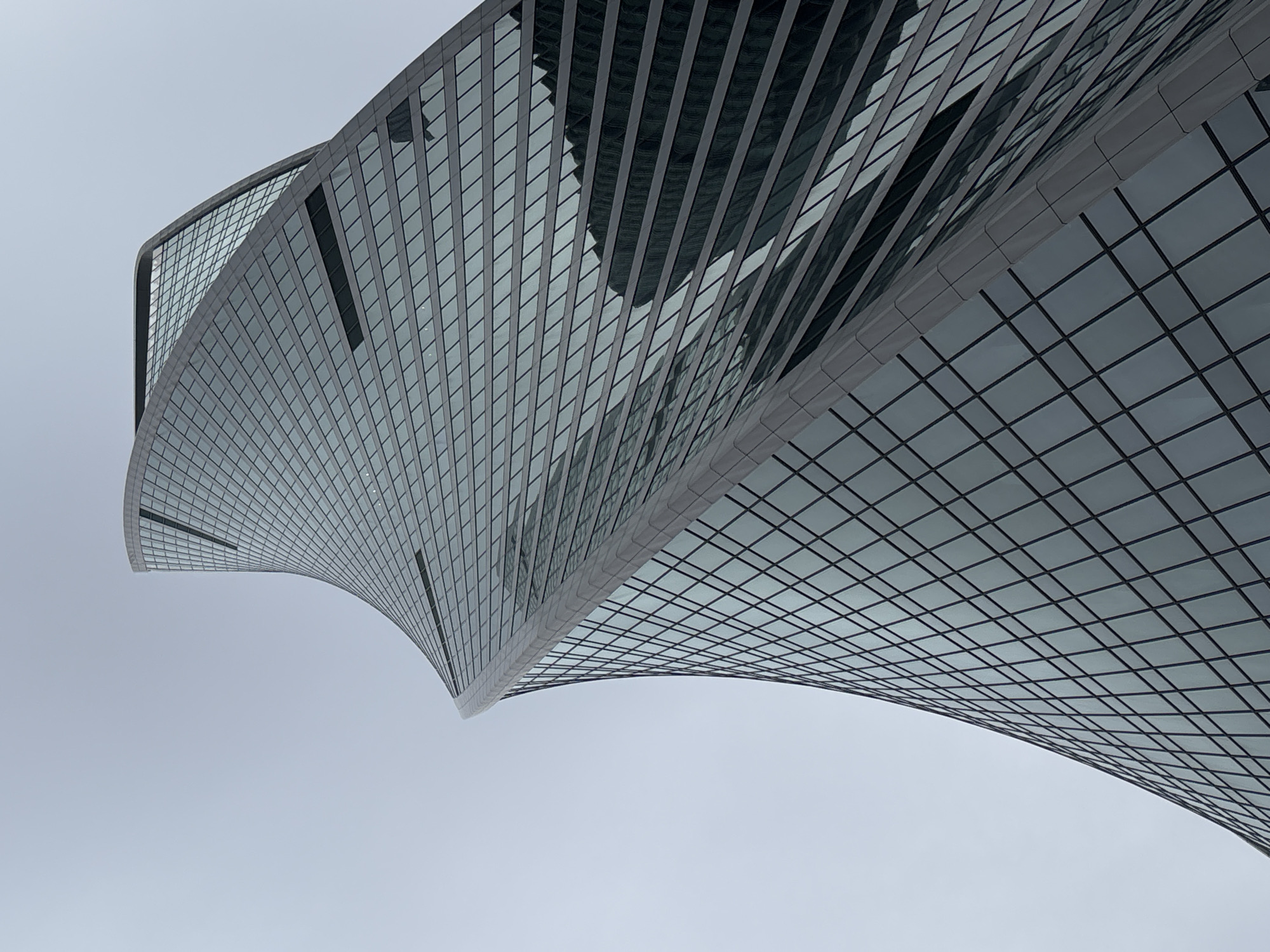 The image depicts a low-angle view of the Evolution Tower, a twisting skyscraper located in the Moscow International Business Center, Russia. The building is primarily constructed of glass panels arranged in a grid-like pattern, reflecting the sky and other surrounding buildings. The tower's unique spiraling design is prominent in the image, showcasing its curved shape. The sky is a uniform light gray, providing a neutral backdrop that emphasizes the architectural details of the tower. There are no people visible in the image.