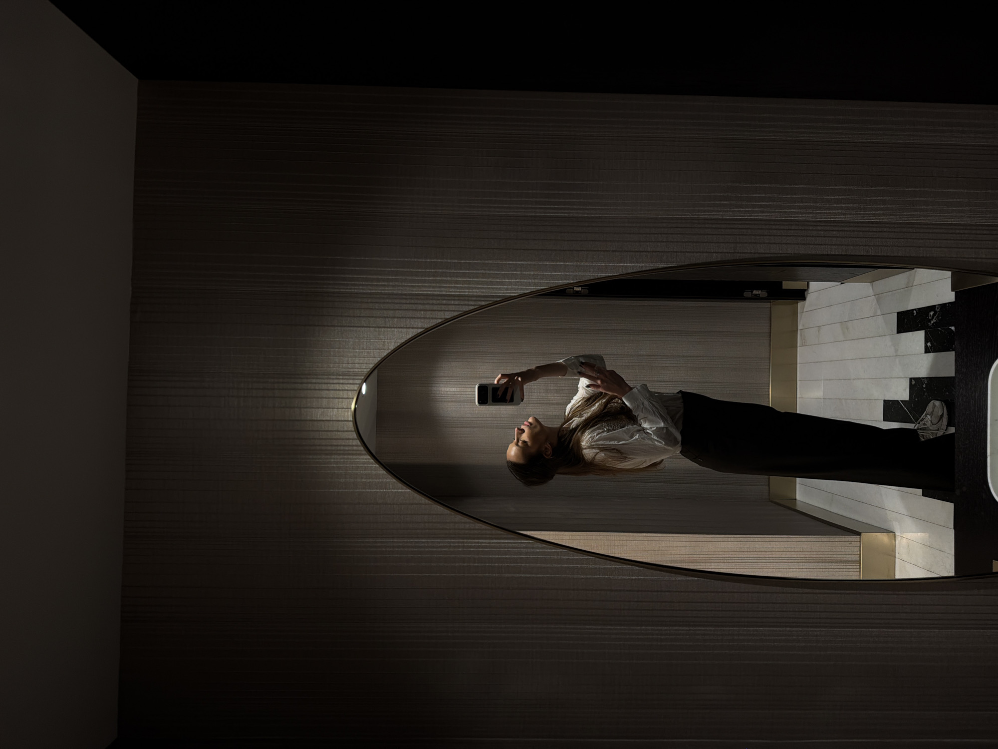 The image shows the reflection of a woman taking a selfie in an oval mirror. The woman is wearing a white long-sleeved shirt and dark pants. She is holding a smartphone in her hand and facing the mirror. The reflection shows the woman standing on a tiled floor with a black and white pattern. The background includes a wall with a dark, textured finish. The location appears to be an indoor space, possibly a bathroom. The scene is well-lit, highlighting the woman and the surrounding environment. The city is mentioned to be Budva, Montenegro.