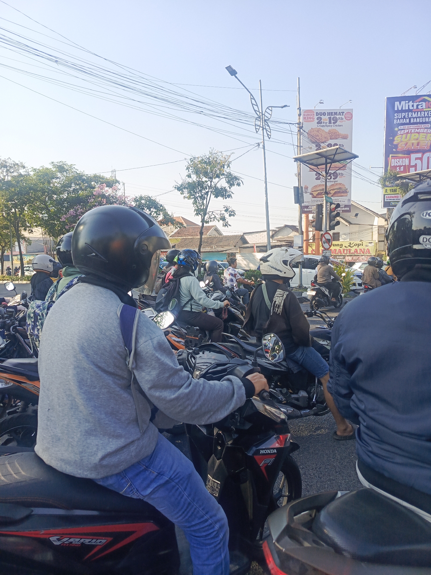 In a city scene in Surabaya, Indonesia, a group of people on motorcycles are stopped in traffic. The sky is clear and blue, with power lines crisscrossing overhead. Trees with pink flowers line the street. Several billboards are visible, one advertising a Burger King promotion and another for a supermarket sale. People are wearing helmets and casual clothing. The motorcycles are parked closely together on the asphalt road.