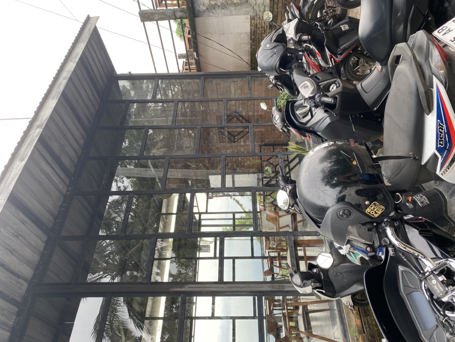 The image shows a view of a building with a large glass facade, in Kecamatan Piyungan, Indonesia. The building is constructed with a corrugated metal roof. The windows are framed in black metal and divided into numerous panes. Behind the building, there is a view of a forested area. The building is empty, but four motorcycles are parked in front of it. The motorcycles are parked along a walkway, and all are facing the street. The motorcycles are black and red, and they are all equipped with helmets. One of the helmets is black and has a sticker on it that reads "KCP 0328". There is a sign on the motorcycle that reads "BEAT" in red. The sign is located on the side of the motorcycle and is written in white. The motorcycle is facing the viewer. The building is located at -7.855 latitude and 110.473 longitude.