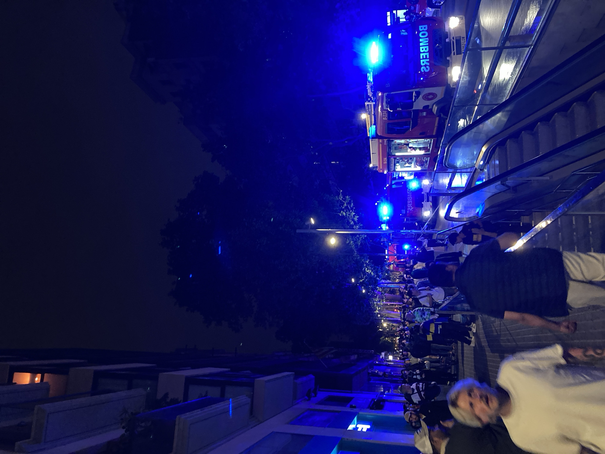 The image shows a street scene at night in an urban area of Unknown, Unknown. A crowd of people is gathered on the sidewalk. Several fire trucks with flashing blue lights are visible. An escalator is positioned near the street, and buildings line the side of the road. Streetlights illuminate the area.