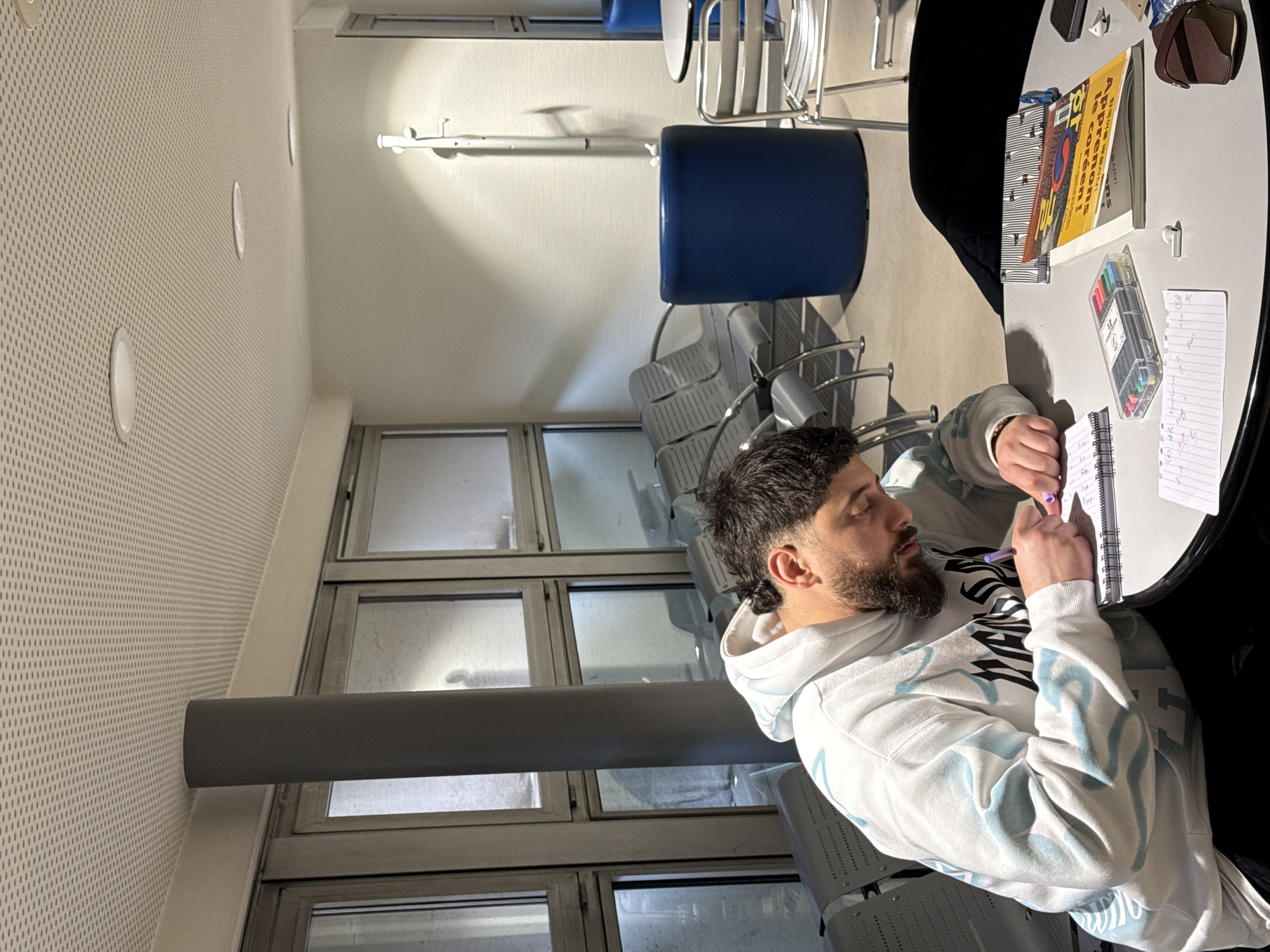A man sits at a table in what appears to be a waiting area or lounge, possibly at the Strasbourg Entzheim Airport in Strasbourg, France. The man, dressed in a white hoodie with black and blue designs, is actively writing in a notebook with a purple pen. On the table, next to the notebook, there are markers, a notebook, and an open book with the visible title "Après nous le Korean!" The environment includes metal framed windows with partial views to the exterior and rows of grey plastic chairs and blue plastic chairs and a coat rack along the wall. The room is well-lit, and the layout suggests a modern, functional design.