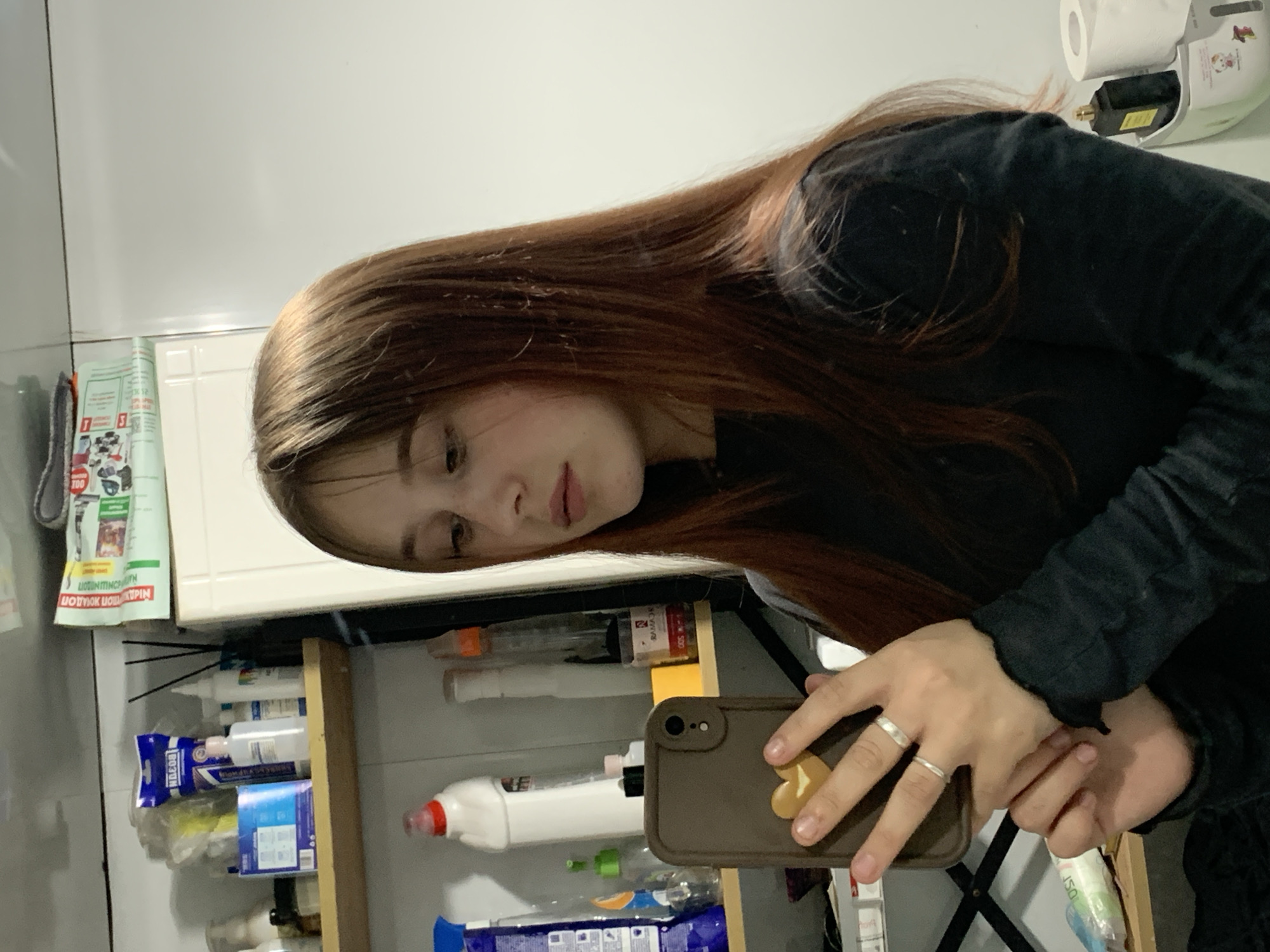 A young woman with long brown hair is taking a selfie in what appears to be a bathroom. She is wearing a black shirt. In her hands, she holds a brown phone case. A cabinet with various cleaning supplies is visible behind her, as well as a roll of toilet paper. The location of Фоки, Russia is irrelevant as there is no information present in the image that confirms that.
