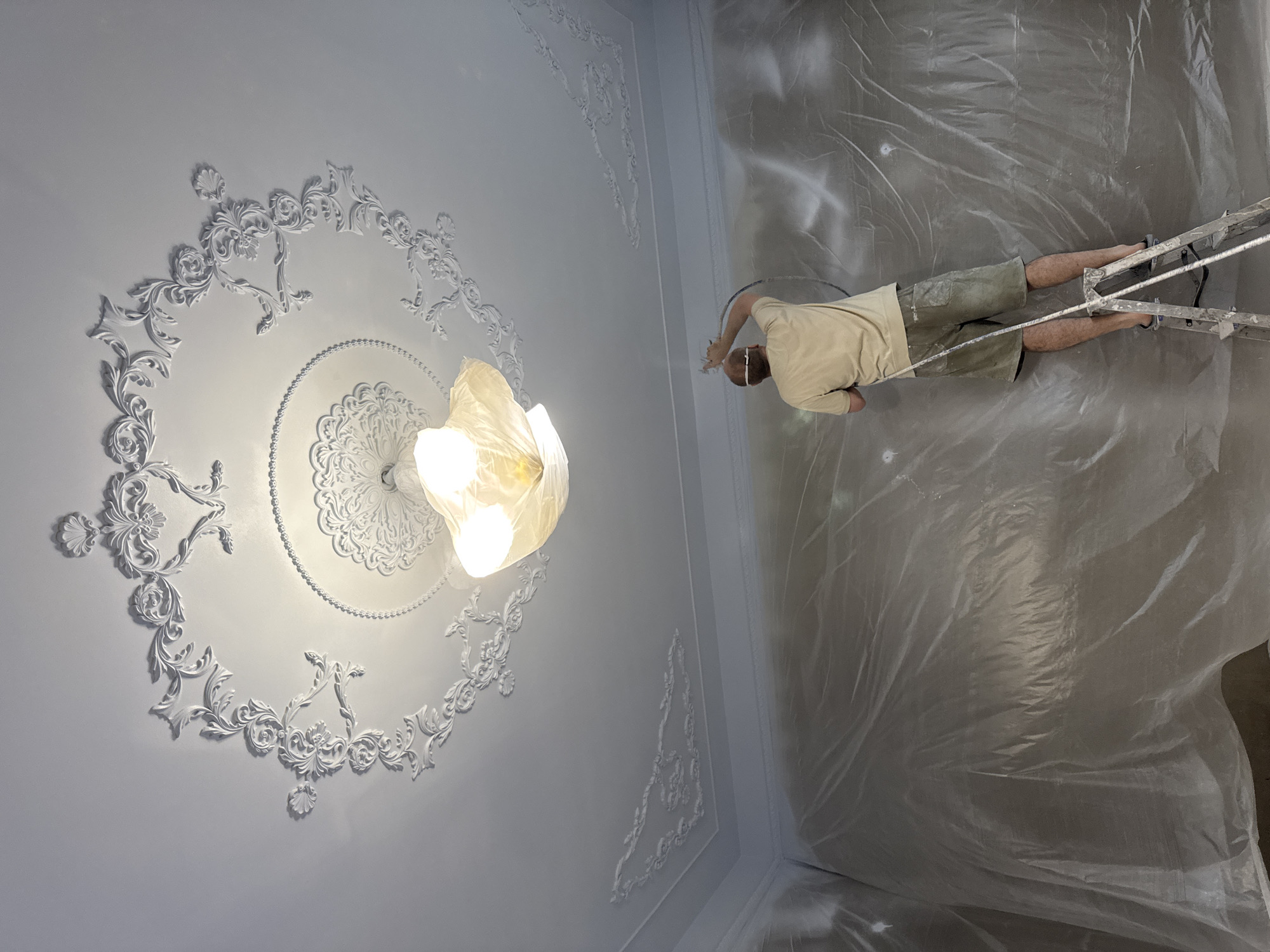 The image shows a man painting the ceiling of a room. The room is covered in plastic sheeting. The ceiling is white and has ornate plasterwork details, including a decorative medallion around a light fixture. The man is standing on a ladder and wearing a mask. He is holding a paint sprayer. The location is likely an interior space in Chaikovsky, Russia, based on the background information.