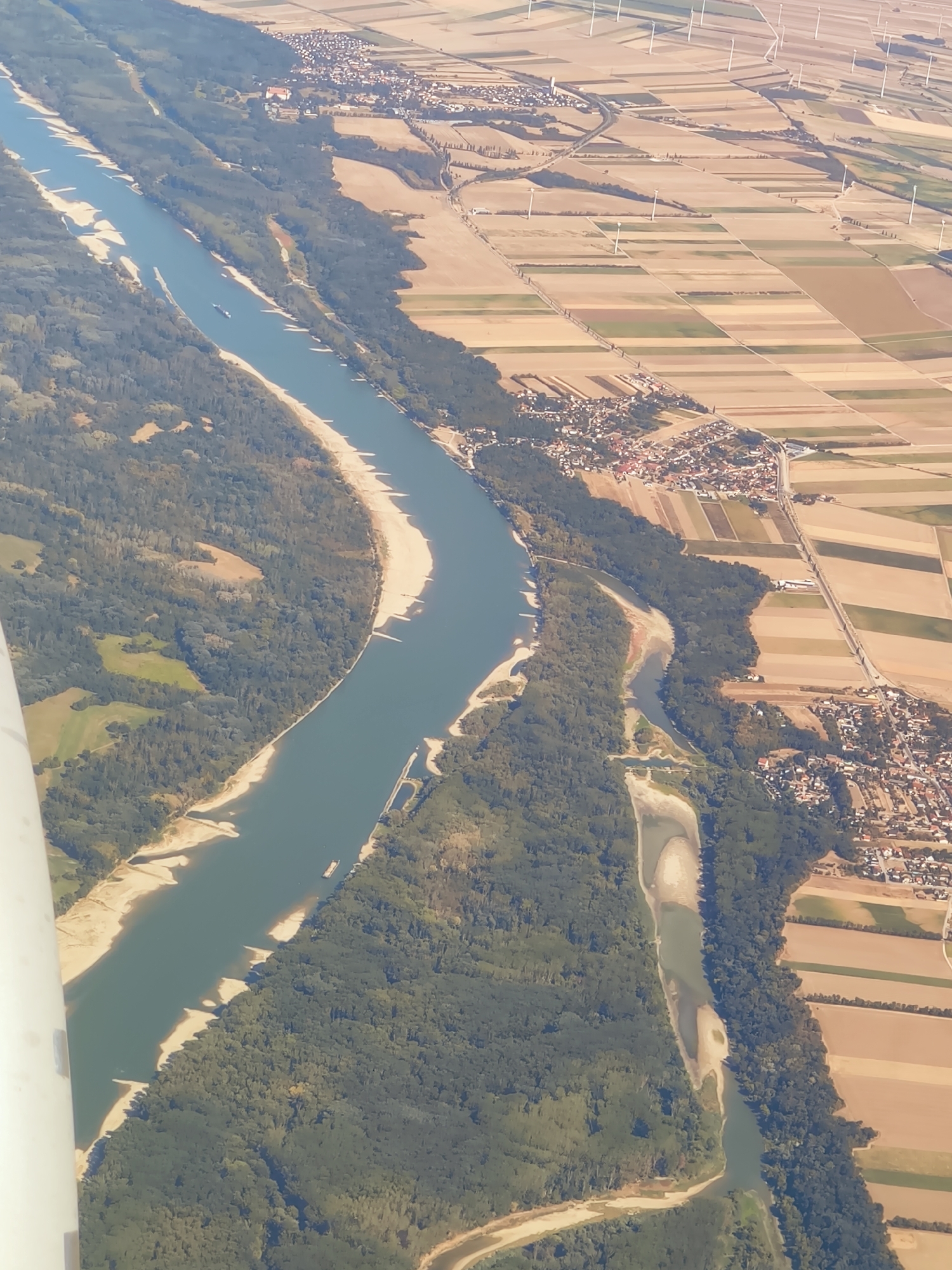 An aerial view captures the Danube River flowing through a landscape near Haslau-Maria Ellend, Austria. The river is wide and greenish-blue, with sandy banks and some exposed sandbars. Dense green forests line the river on the left side of the image, with patches of lighter green and brown fields interspersed. On the right side of the river, agricultural fields are visible, laid out in a patchwork of brown, tan, and green. Numerous wind turbines are scattered across the fields in the background. A small town or village with red-tiled roofs is situated along the riverbank, adjacent to the fields. A barge is visible on the river. The foreground of the image is partially obscured by a white object, likely part of the aircraft from which the photo was taken. The lighting suggests it is daytime, with the sun casting shadows.