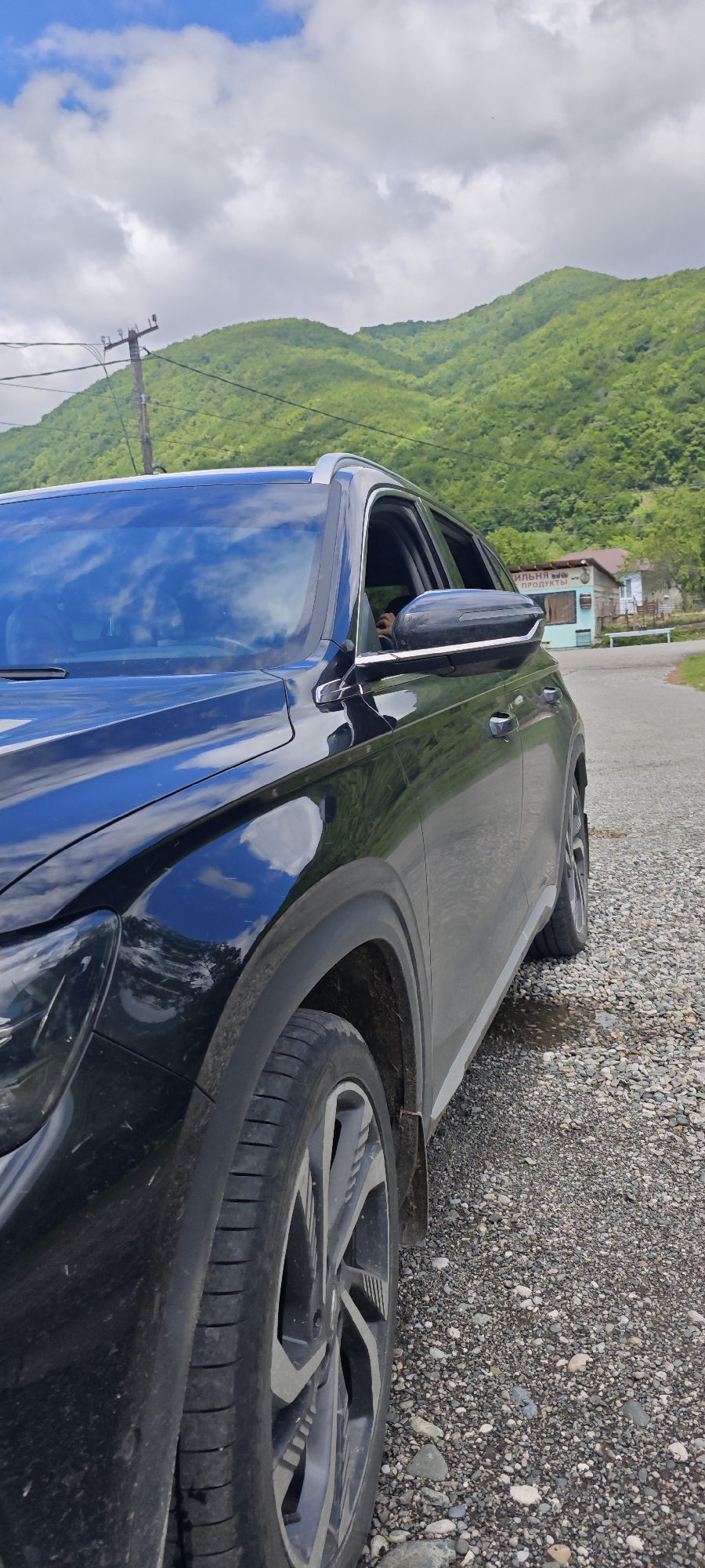 A black SUV or crossover is prominently featured, parked on a gravel shoulder beside an asphalt road. The vehicle is angled towards the right, showcasing its front passenger side. The glossy black paint and windshield reflect the bright, partly cloudy sky and the verdant mountains. A person's hand or arm is visible through the slightly lowered front passenger window.
The scene is set in a mountainous and rural landscape, specifically Bzypta, Abkhazia. Lush green, tree-covered mountains rise in the background under a sky dotted with white clouds and patches of blue. In the mid-ground on the right, a small, light-colored building with a brown roof, appearing to be a local shop or kiosk, stands near the road. Visible Cyrillic text on its facade includes "ПРОДУКТЫ" (PRODUKTY), indicating "GROCERIES" or "PRODUCTS." A utility pole with wires is present on the left, extending towards the hills. The asphalt road curves out of view to the right. The image depicts a daytime scene, likely during late morning or afternoon, suggesting a stop on a scenic journey through this natural environment.