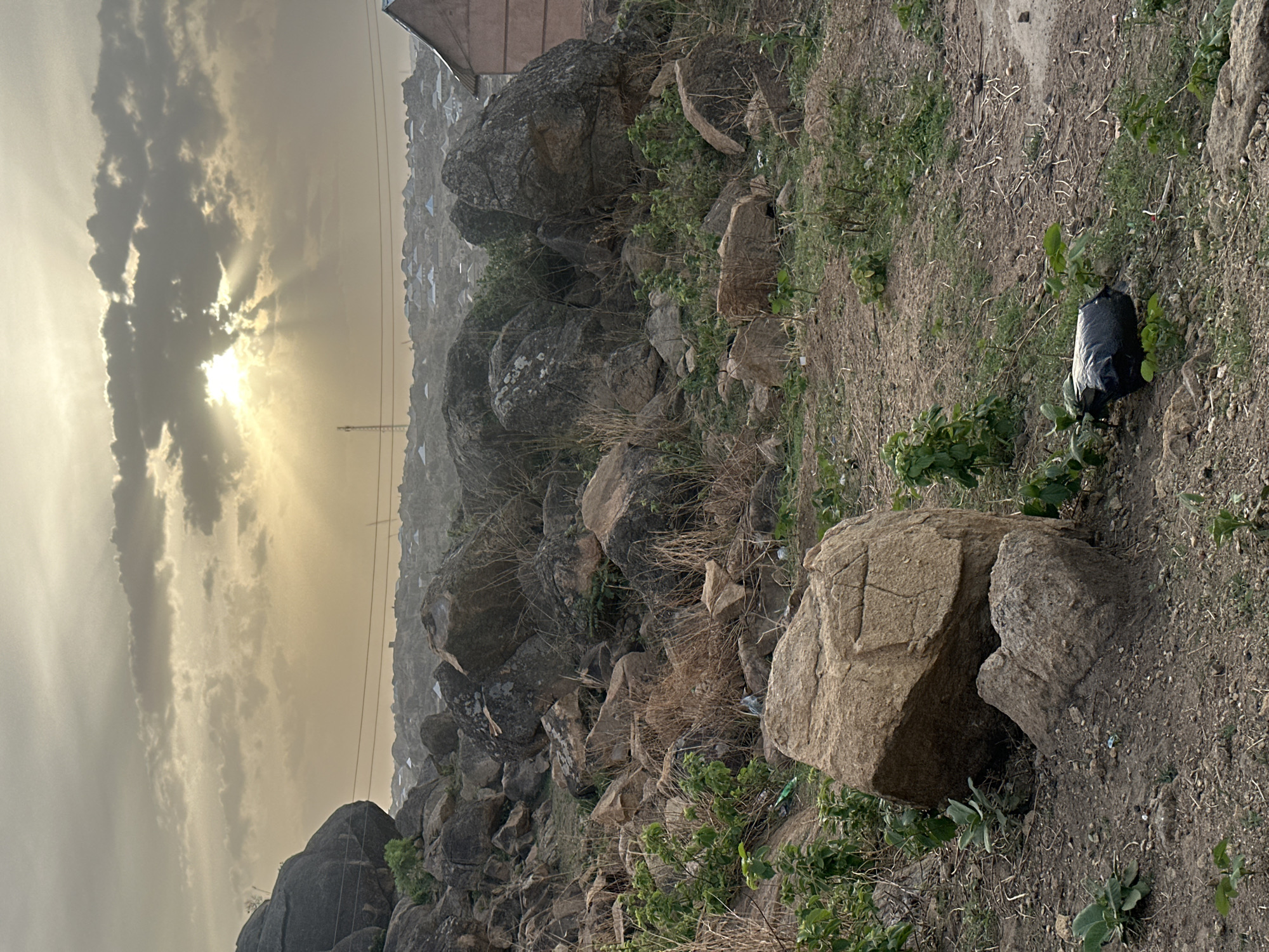 The image shows a rocky landscape in the city of Jos, Nigeria, with the sun obscured by clouds and emitting rays of light. There are houses in the background, and a black bag is sitting on the ground among rocks and sparse vegetation. A large boulder is in the foreground. The scene is captured from a slightly elevated perspective.