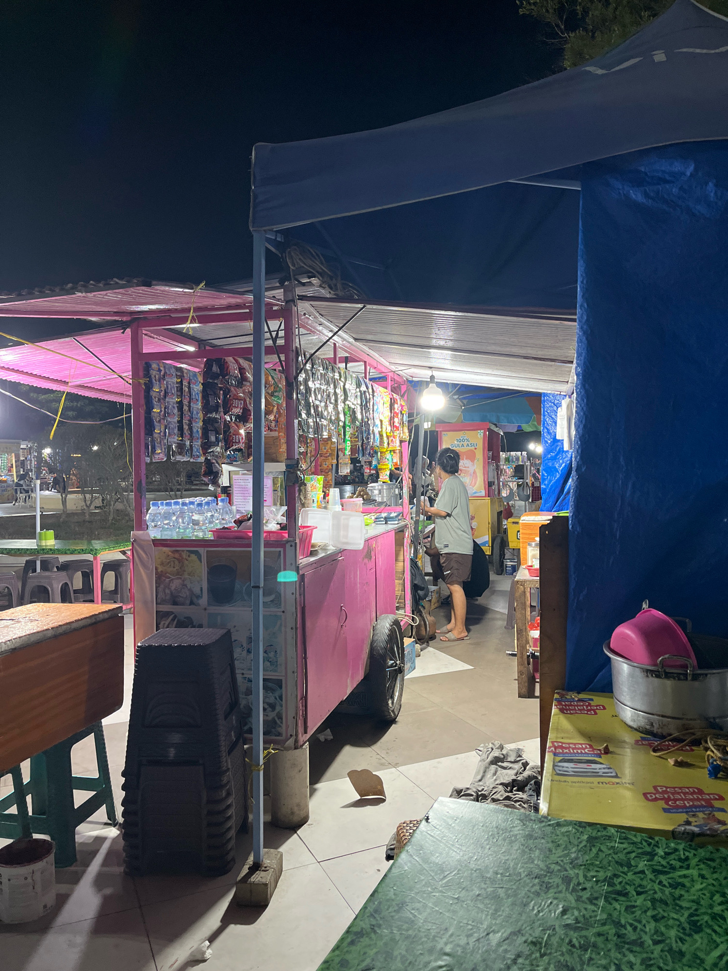 This image depicts a nighttime scene at a street food stall in Kendari, Indonesia. The stall is brightly lit by overhead lights, illuminating a variety of goods, including packaged snacks hanging from racks and bottles of water. A person, wearing a light-colored t-shirt and shorts, is standing behind the counter, appearing to be attending to customers or preparing food. The stall itself is colorful, primarily pink and blue, with various signs and posters displayed. In the foreground, there are stacks of plastic stools and a table covered with what appears to be a green grass-patterned cloth. The ground is tiled. The background shows more stalls and trees, suggesting a bustling market or street food area.