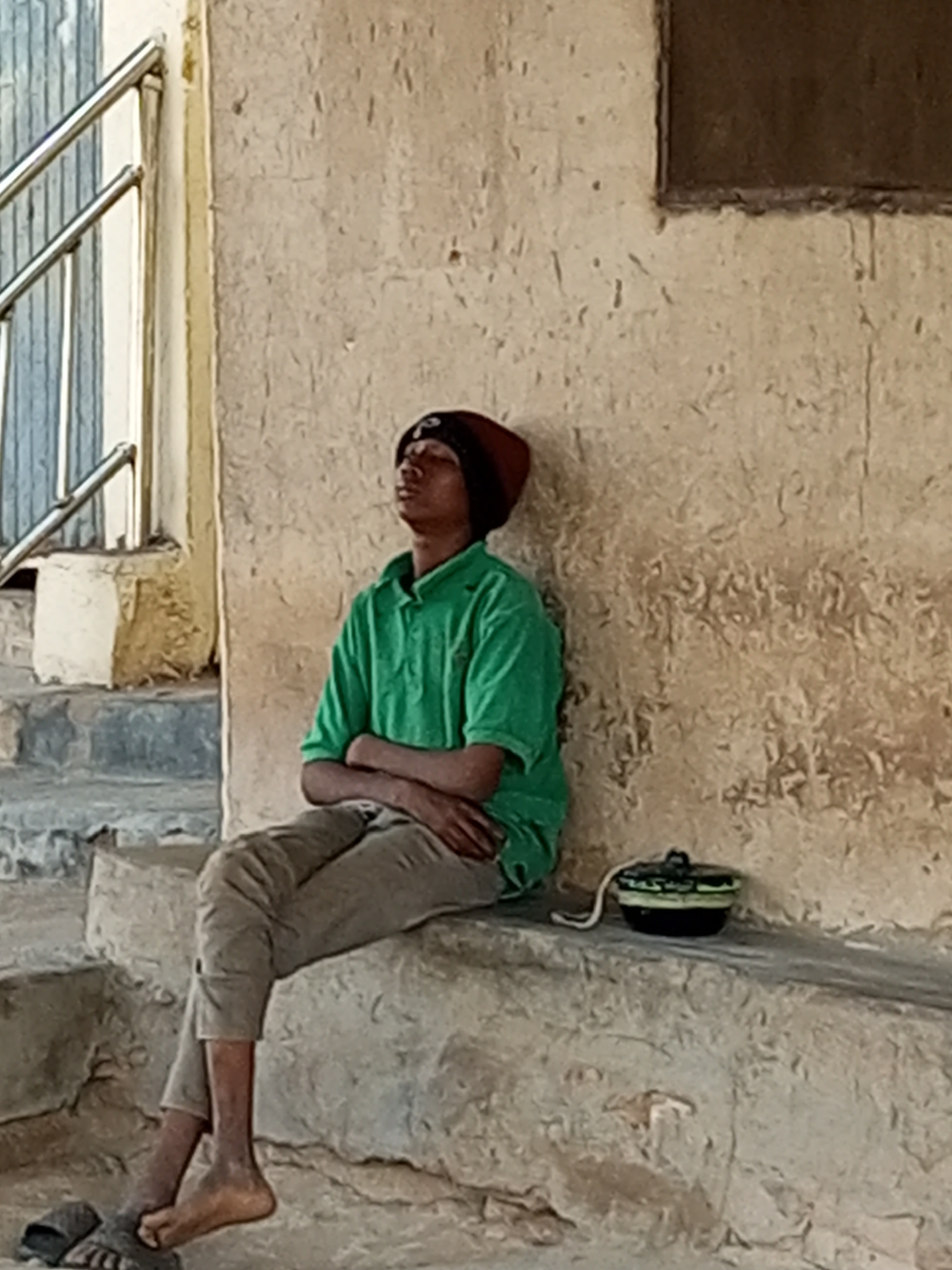 In Arawa, Nigeria, a young male with dark skin, wearing a maroon beanie and a green polo shirt, sits with his arms crossed on a concrete ledge. His eyes are closed, suggesting he is resting or contemplating. He is wearing light brown pants and dark sandals. Beside him on the ledge sits a small black container with a white string. The background consists of a textured, light-colored wall and some steps with a metal railing.