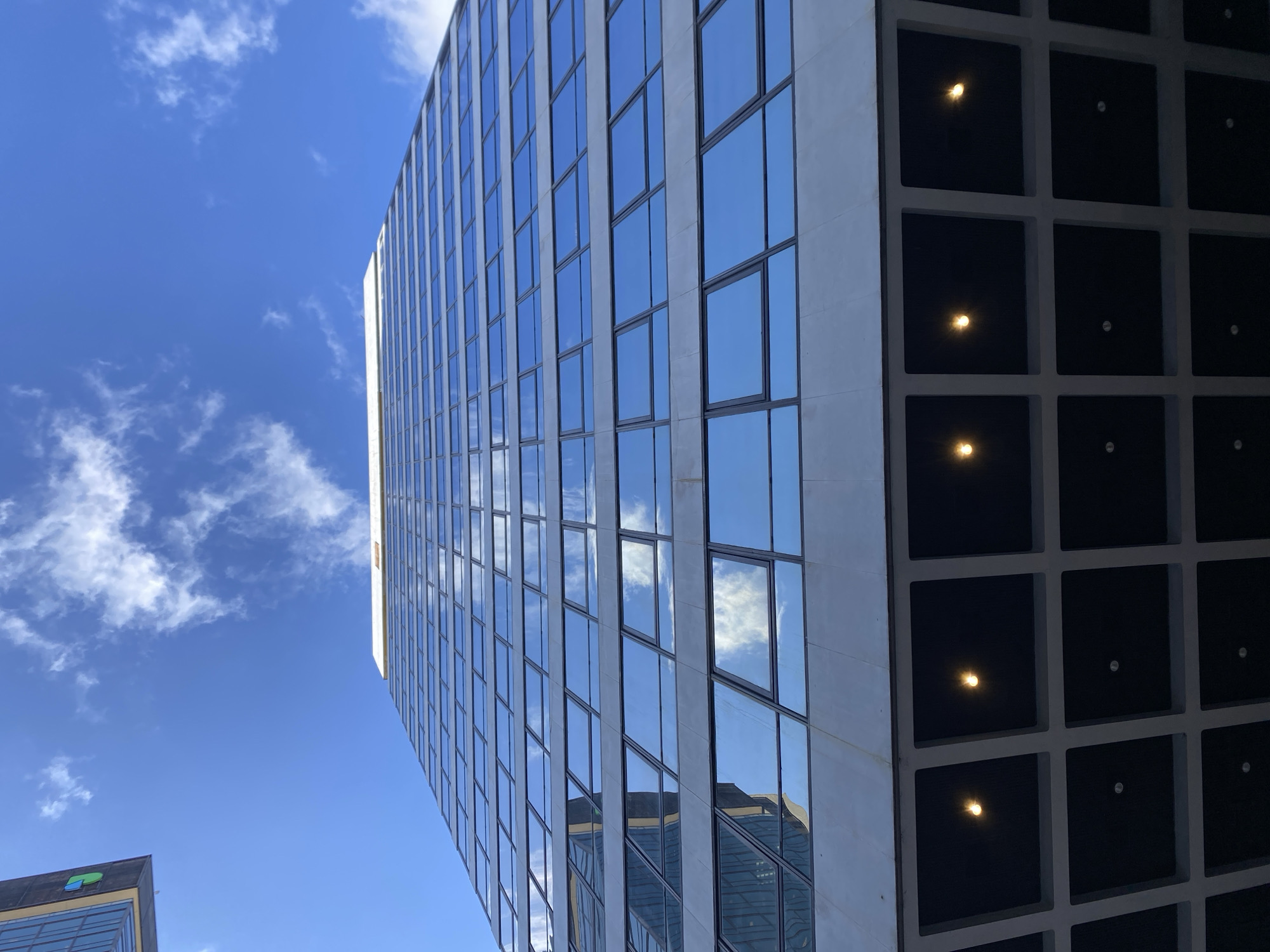 The image presents a striking upward view of modern urban architecture under a bright blue sky. A prominent high-rise building dominates the frame, showcasing a sleek, blue-tinted glass facade on its left side. This extensive grid of windows reflects the vibrant blue sky and scattered white clouds, with deeper reflections of another building visible in the lower panes. The window frames are a light, possibly white or light gray, material.
To the right of this reflective facade, the building transitions to a section characterized by a darker, grid-like pattern of recessed square openings. These dark squares appear to be part of an adjacent structural element, possibly a parking garage or service core. Several warm, yellow-orange lights are illuminated within these squares, creating a visual contrast with the darker background.
In the lower-left corner, the top edge of a second building is partially visible. This structure appears to have a darker, possibly gray or black, exterior and features a distinctive green and blue logo on its roofline. The logo is a stylized symbol, though any associated text is not discernible.
The background is a clear, brilliant blue sky adorned with fluffy white cumulus clouds, indicating a daytime setting with good weather. The image captures the interplay of light, reflection, and modern design, with no human activity visible. No readable text is present within the scene.