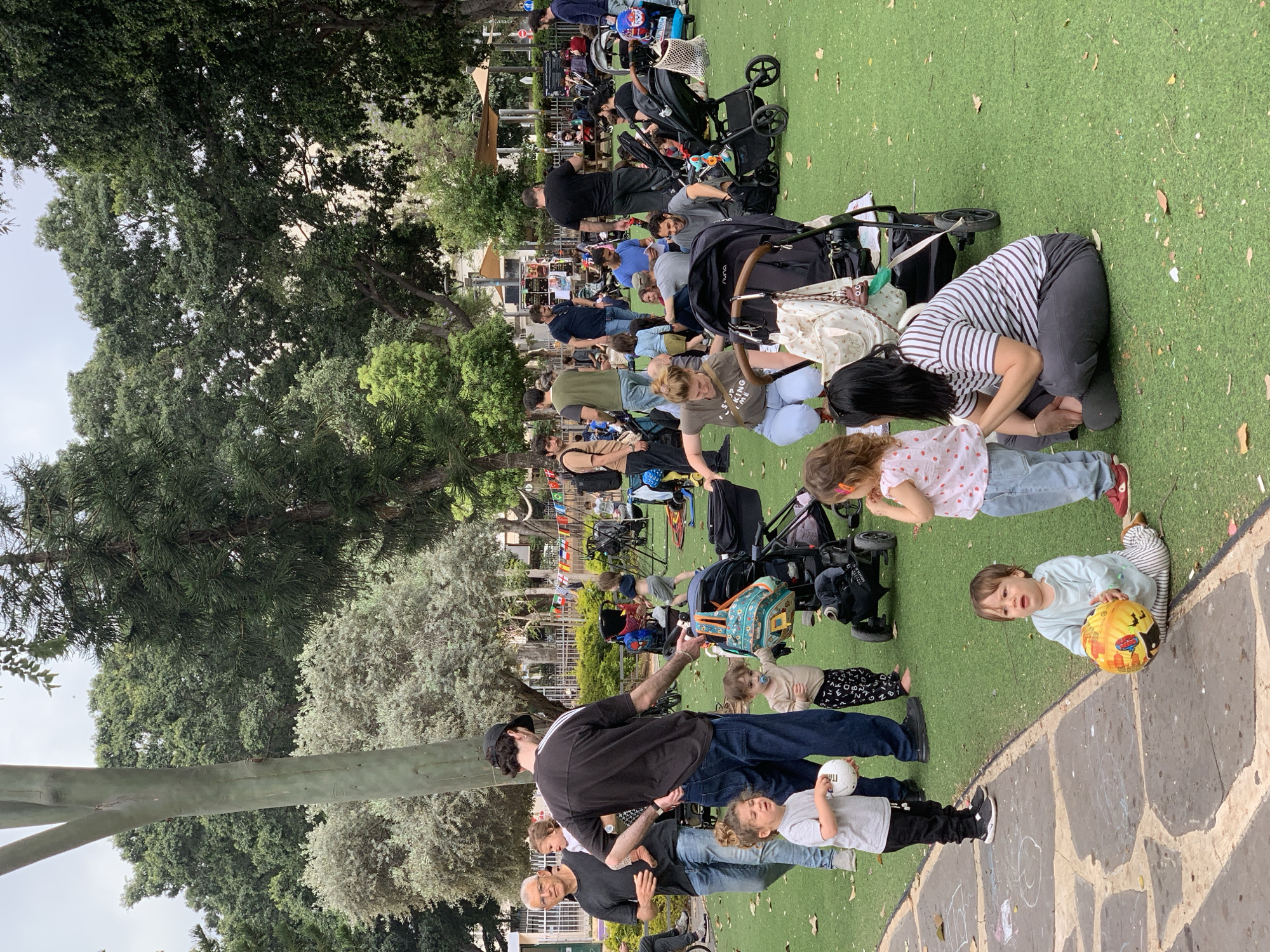 The image depicts a gathering of people in a park, likely parents and young children, situated on artificial turf. In the background, a row of buildings is visible beyond a fenced area with colorful flags strung up, possibly marking a playground or communal space. Various strollers are scattered among the people. Several individuals are sitting or kneeling on the ground, attending to the children. In the foreground, there are several children, one of whom is holding a yellow ball. The location is in Tel-Aviv, Israel.
