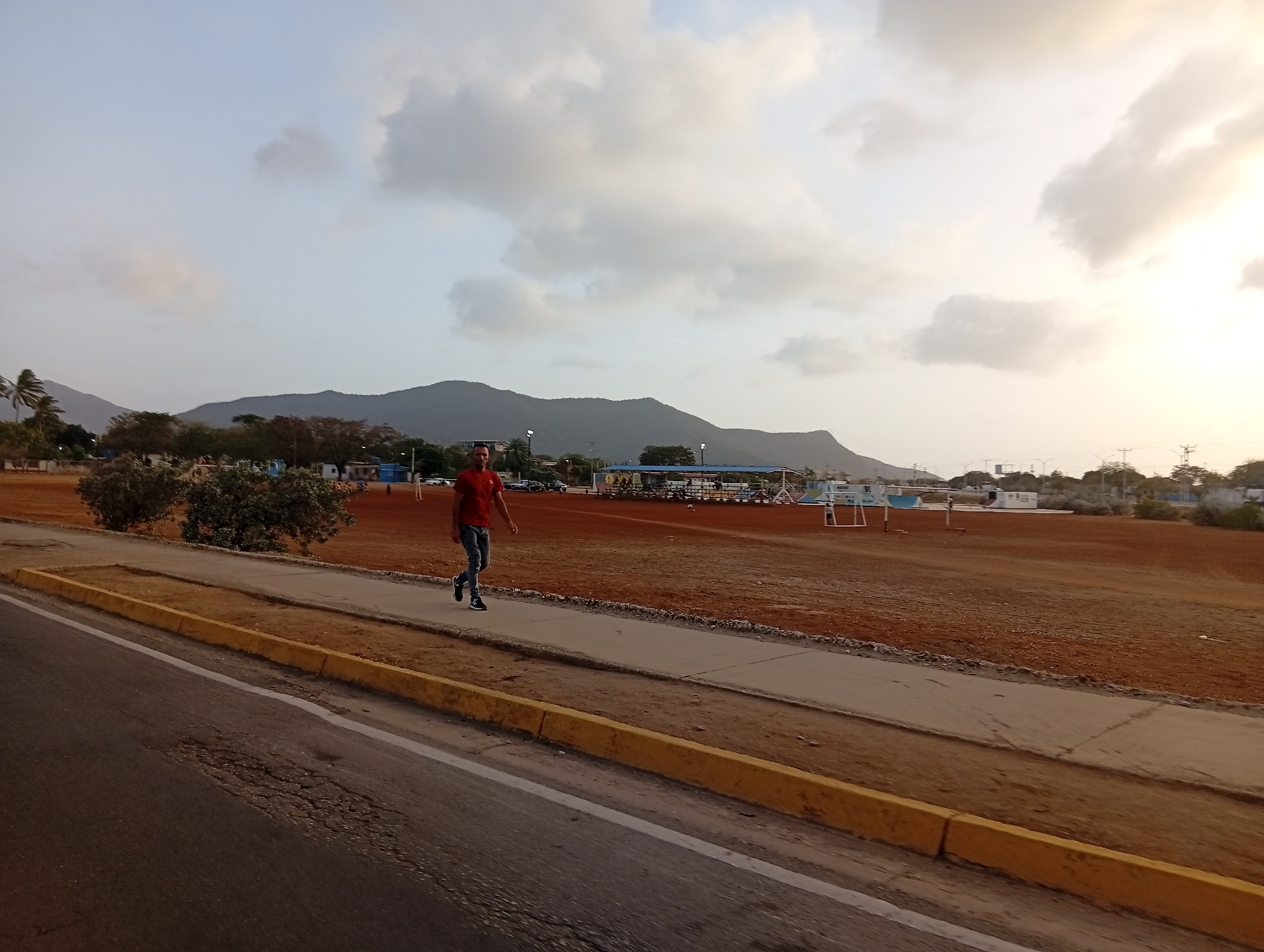 The image captures a view in Juan Griego, Venezuela, featuring a man walking along a sidewalk next to a road. The road has a noticeable patched area, and a yellow curb separates it from the sidewalk. Beyond the sidewalk, there's a large, reddish-brown open field that seems to be a sports ground, identifiable by soccer goalposts in the distance. In the background, mountains rise, and the sky is filled with clouds tinged with the colors of the setting sun. Some trees are also scattered near the edge of the field.
