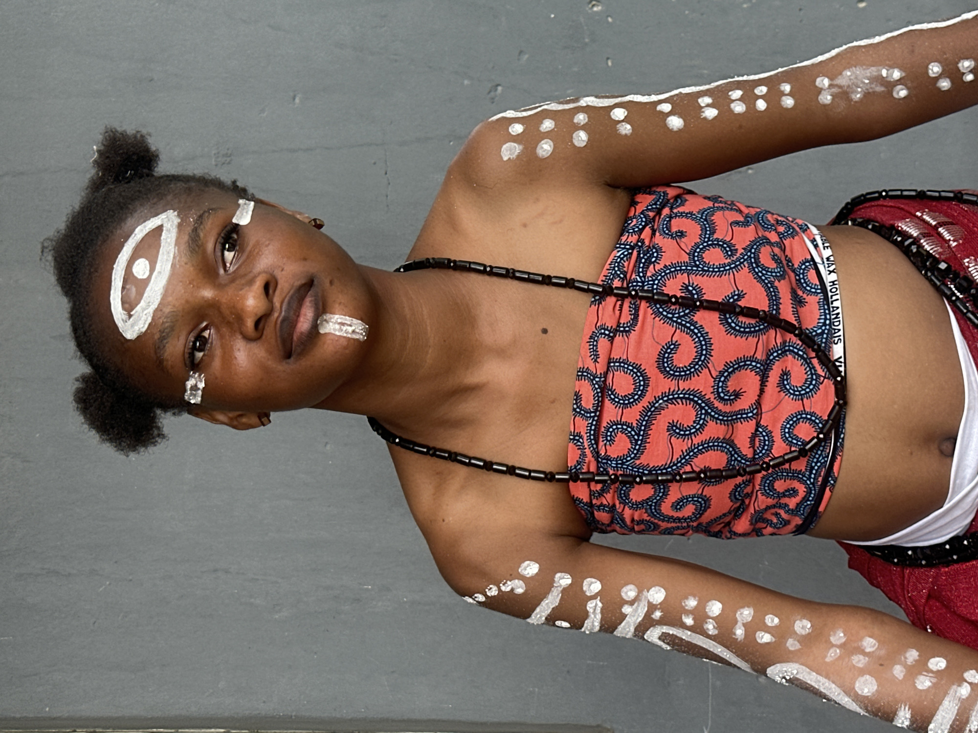 In an outdoor setting against a gray, concrete wall, a young Black woman is depicted in a medium shot, looking to her right with a slight head tilt. She has dark hair styled in two small buns atop her head. Her face is adorned with white face paint, featuring an eye-like design on her forehead, two rectangular markings below her eyes, and a vertical stripe on her lower lip. She wears a strapless, coral-colored tube top with a repeating blue, swirling pattern. A dark, beaded necklace rests on her chest. Her arms are also decorated with white paint, showing abstract patterns and dots. A glimpse of a red garment with reflective sequins is visible below her tube top, and a white sash is tied around her waist. The image captures the woman in a posed, possibly ceremonial, context. This scene is located in Badagry, Nigeria.