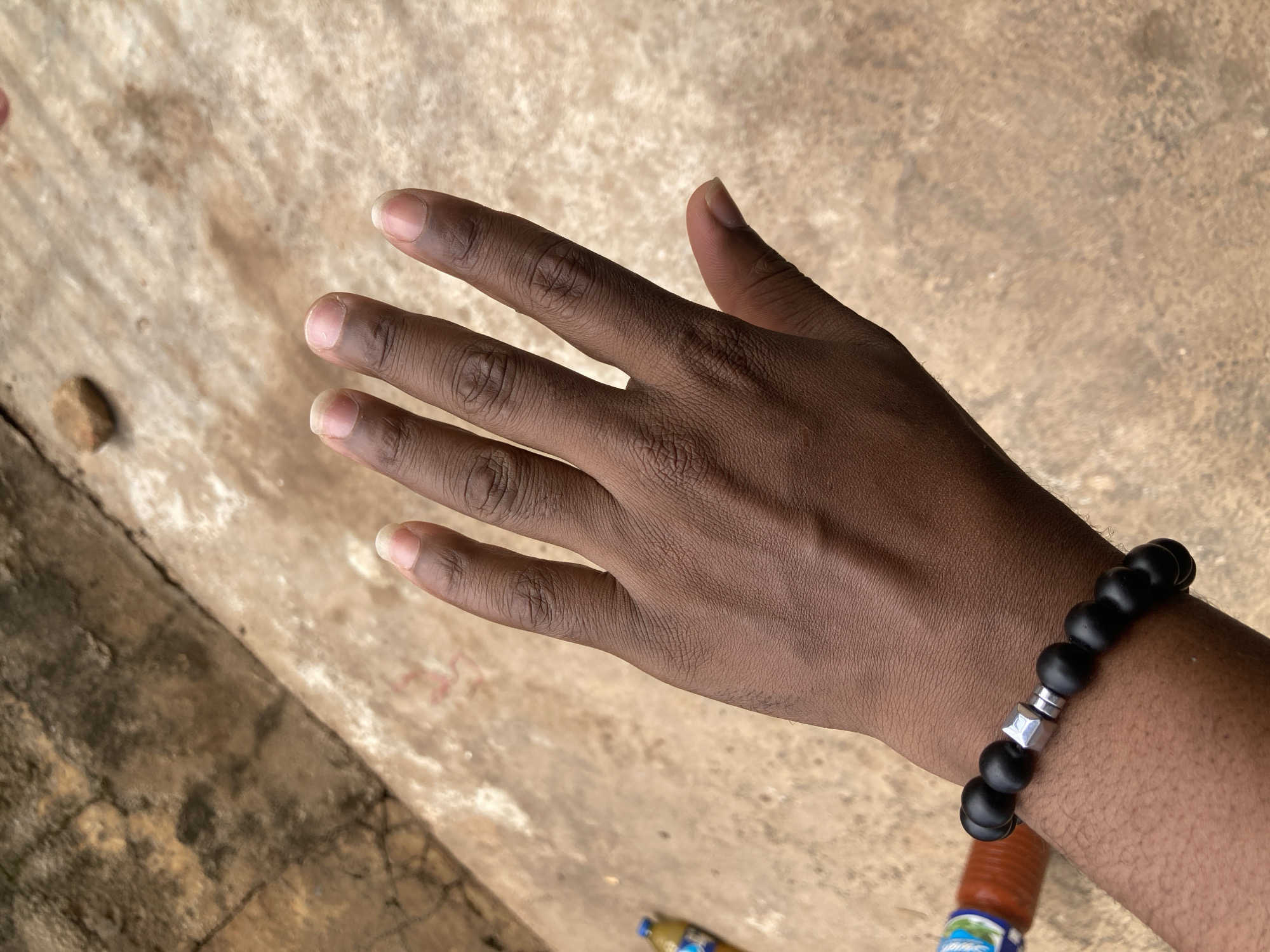 A person's left hand, palm facing upwards, is prominently displayed against a textured, light brown concrete surface. The hand exhibits a dark skin tone, with visible veins and skin creases. The fingernails are short and unpolished. The person is wearing a black beaded bracelet with a silver square-shaped charm on the wrist. In the background, out of focus, is a blurry orange bottle with a blue and white label, lying on its side on the concrete. The scene is illuminated by natural light, casting subtle shadows. The concrete surface appears weathered and slightly uneven, with some darker, possibly damp patches and a small, brown pebble to the upper left of the hand. The overall impression is an outdoor setting, possibly in a public area or courtyard in Gwong, Nigeria.