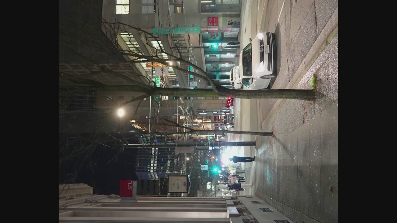 The image depicts a nighttime street scene in Vancouver, Canada, likely on Granville Street, judging by the "Granville St" street sign visible. A person stands on the sidewalk, looking towards the street. Several other pedestrians walk along the sidewalk. A white Volkswagen Jetta drives down the street, its headlights illuminated. The street is wet, indicating recent rain. Trees with bare branches are planted along the sidewalk. Buildings with illuminated windows line the street. A red SFU (Simon Fraser University) sign is visible on a building facade. A clock hangs on a pole near the sidewalk. The overall atmosphere is urban and nocturnal.