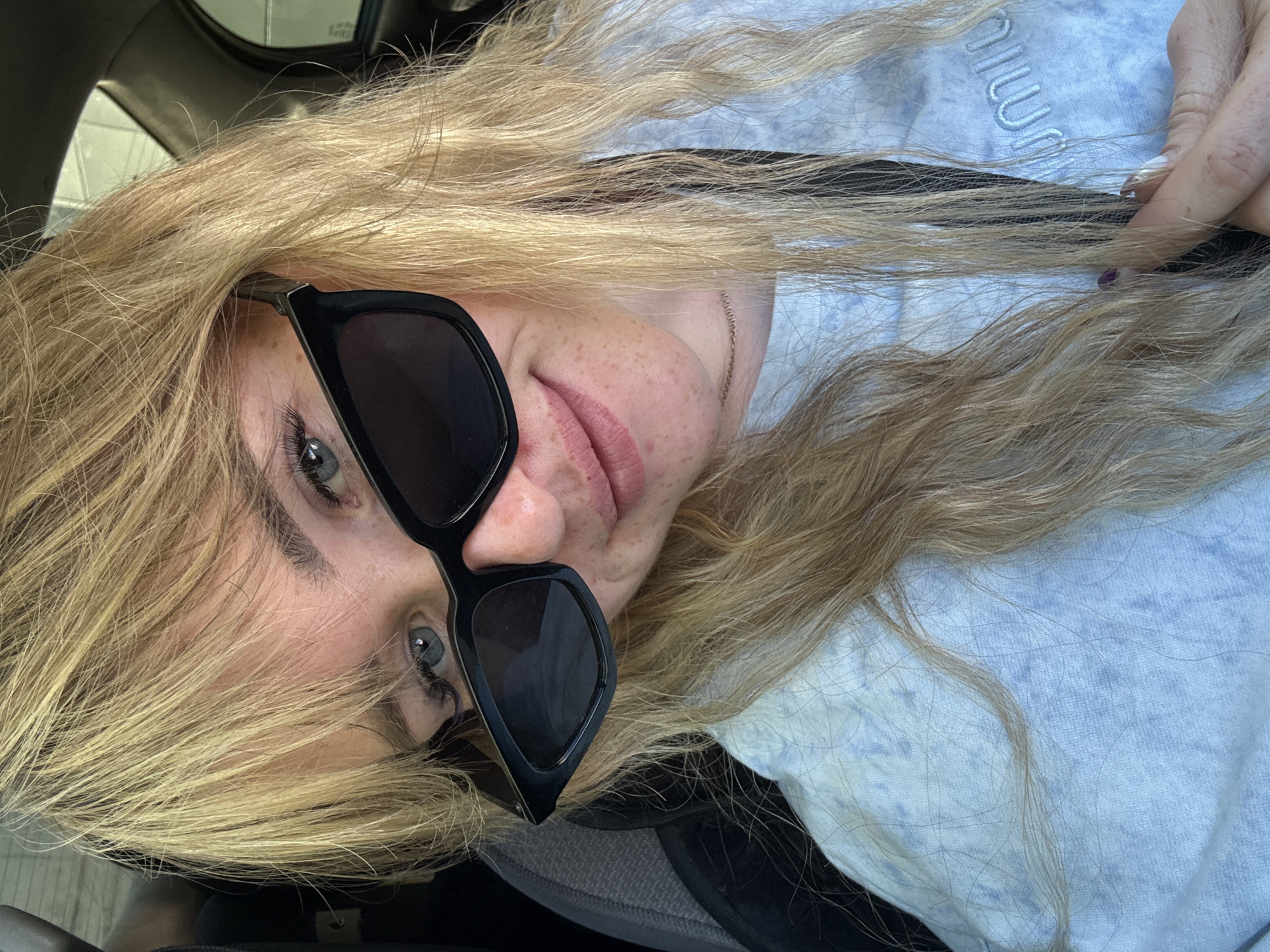 A young woman with blonde hair and blue eyes is pictured from the chest up, wearing black sunglasses and a light blue tie-dye t-shirt. She is in a car, with the interior visible in the background. Her hair is partially covering her face and shoulders. She is looking towards the camera with a slight smile. Her left hand is visible, holding onto the t-shirt near her chest. The image is taken from a slightly high angle, looking down at the woman. The lighting suggests it is daytime, with sunlight filtering in. There are no other people visible in the frame. The environment appears to be the interior of a vehicle.