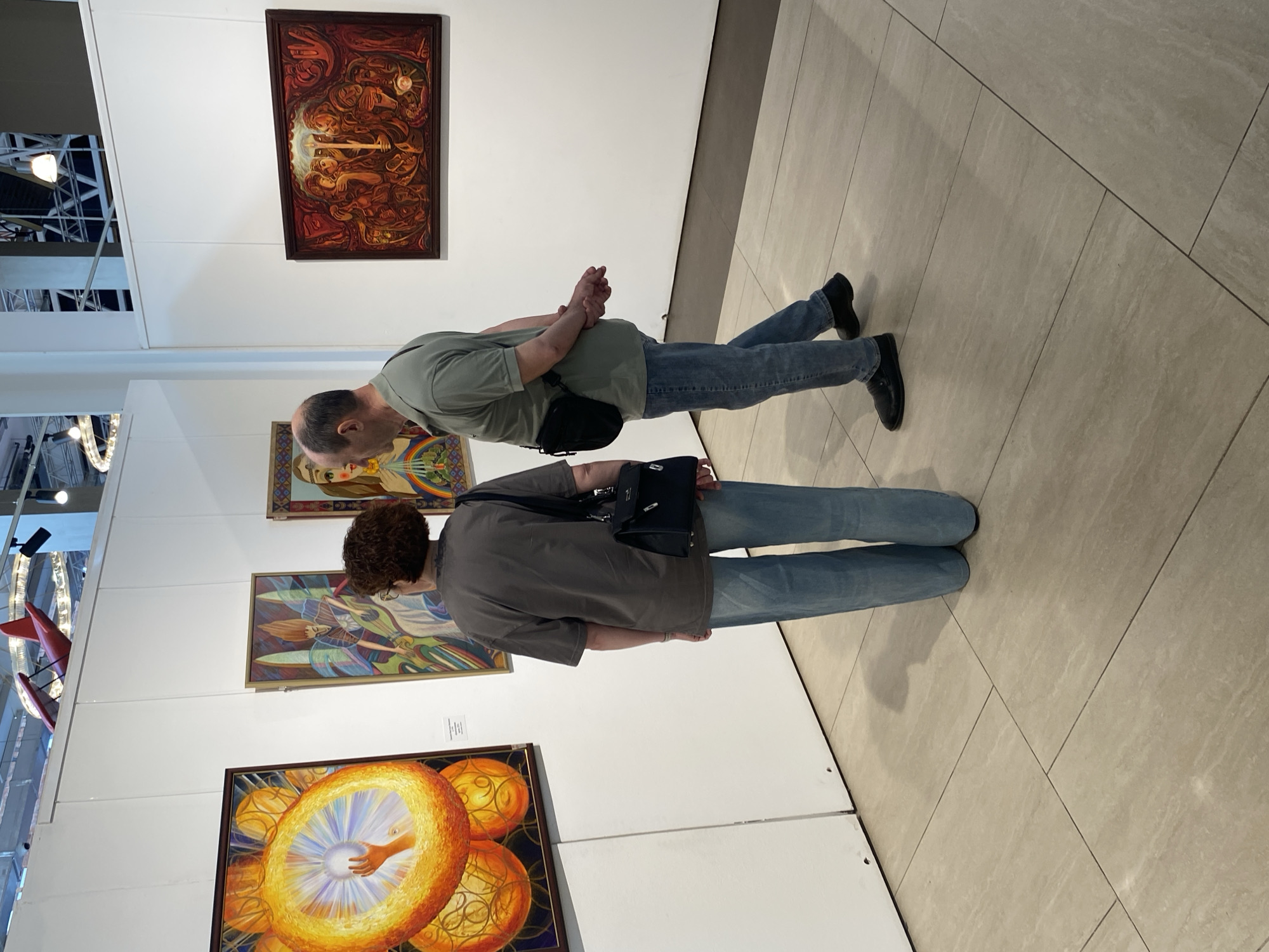 Two individuals stand viewing artwork at an exhibition. The location appears to be an art gallery. Specifically, the scene is at the Yekaterinburg Museum of Fine Arts in Russia. The gallery features white walls, tiled flooring, and displays paintings framed and mounted. The paintings feature a range of styles from figurative art to symbolic or allegorical pieces. The viewer closer to the camera is a woman with curly brown hair and a gray shirt. She is wearing jeans and a dark crossbody bag. A man with a receding hairline stands next to her. He is wearing a light green shirt, jeans, and black shoes. He has a small black bag. The two people are both facing the paintings, in what appears to be a thoughtful and appreciative manner. Overhead structures are visible through glass at the top of the frame, possibly part of the building's architecture.