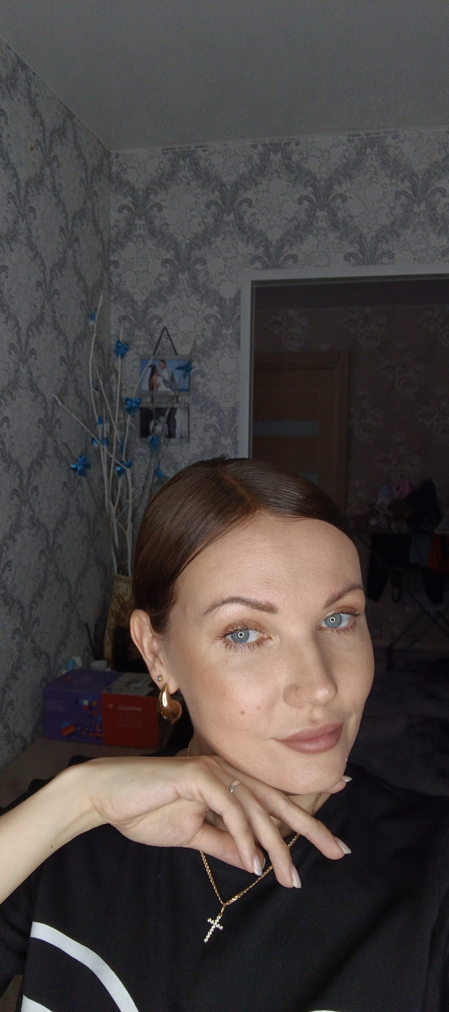 A woman is indoors. She has fair skin, brown hair pulled back, light blue eyes, and wears gold earrings and a gold cross necklace. She is resting her chin on her right hand, which has a silver ring. She wears a black shirt with a white stripe.
The background shows patterned wallpaper and a doorway leading to another room. A decorative item with white branches and blue bows is near a framed photo on a table. The location is in a domestic setting, possibly a residence in the city of Chaikovsky, Russia.