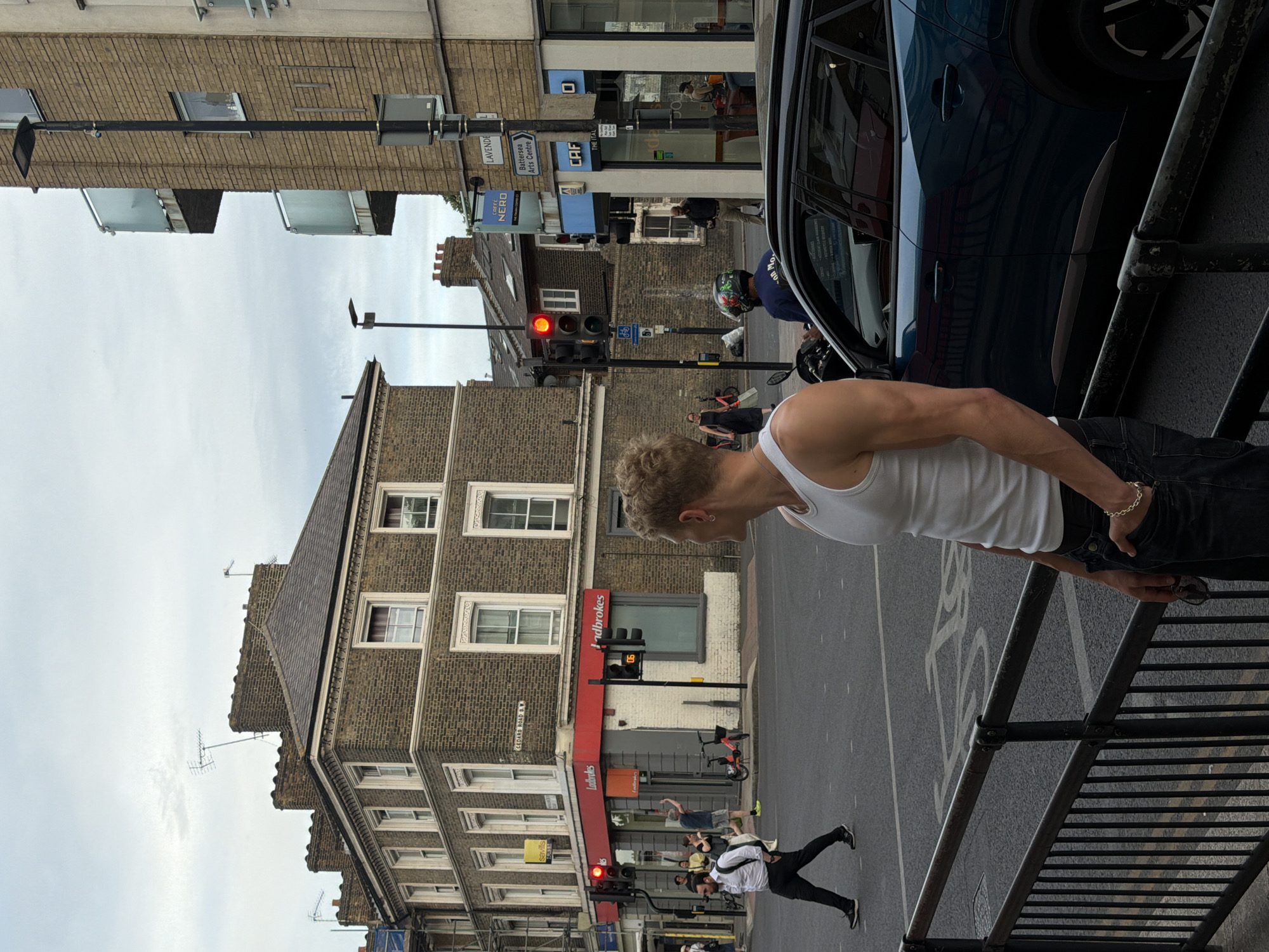 A full-shot medium urban street scene captures a young man in a white tank top and dark jeans standing on a sidewalk in London, United Kingdom. He is seen from the right side, with his head turned to the left. The man has fair skin, blonde hair, and is wearing a silver necklace and a bracelet on his left wrist. He is holding a pair of sunglasses in his right hand, with his hand in his jeans pocket. To the left of the man, a blue car is parked.
The street corner is busy with pedestrian and vehicular activity. A traffic light is visible, showing a red signal. Pedestrians are crossing the street, and a person on a motorcycle can be seen in the background. The buildings lining the street are made of brick and have multiple stories. One building features a red awning with "Ladbrokes" signage. The sky is overcast. The overall impression is a typical street view in a bustling city.
