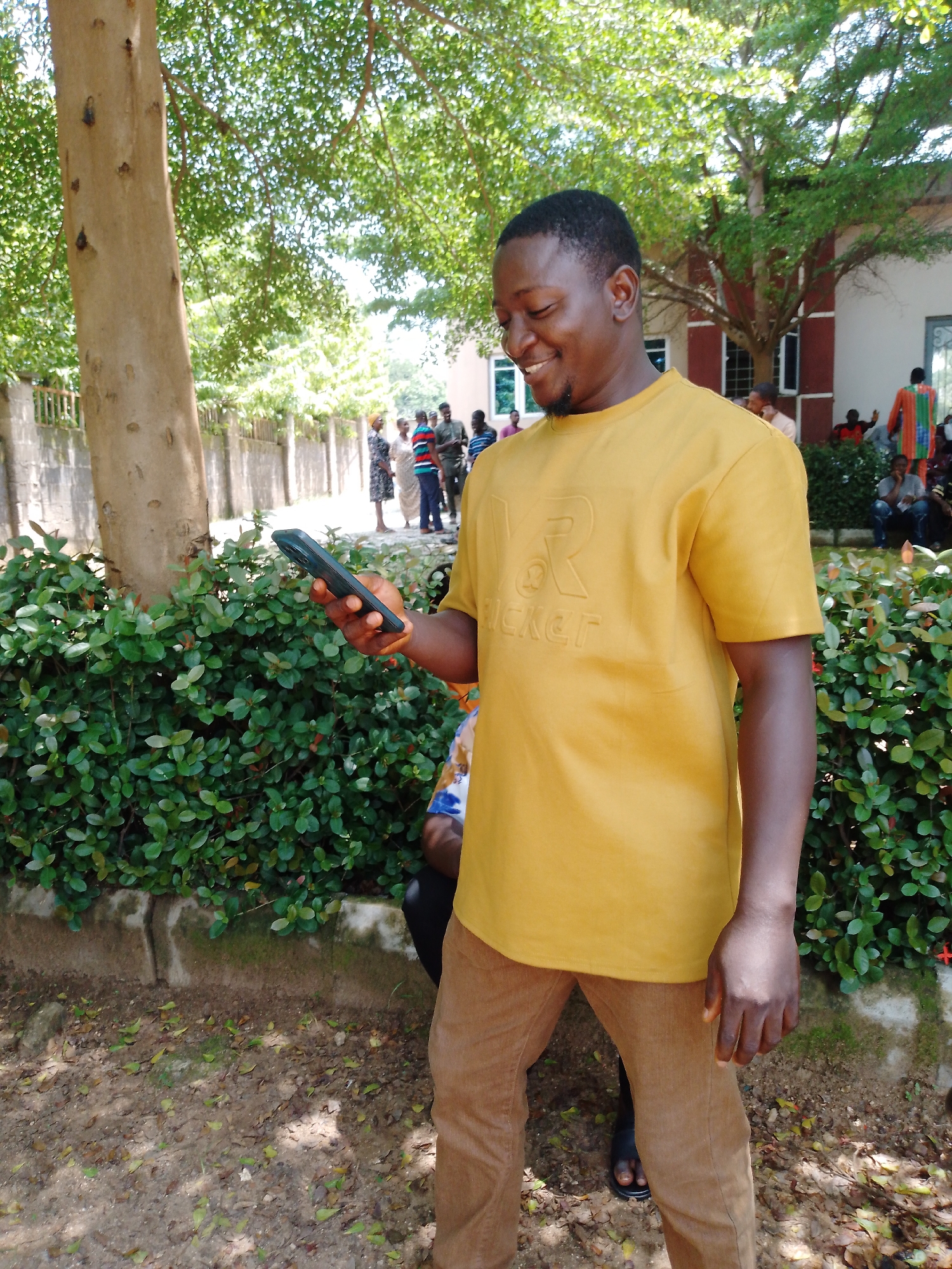 A man in a yellow t-shirt and brown pants is holding a dark-colored smartphone in his left hand. He is smiling and looking down at the phone, suggesting he is engaged with its content. The man is outdoors, with a large tree trunk and lush green foliage behind him. In the background, several other people are visible, some standing and others sitting. One person is wearing a vibrantly colored garment. The setting appears to be a public area, possibly a park or an outdoor event, given the presence of multiple individuals and the natural surroundings. The time of day is likely daytime, as evidenced by the bright sunlight filtering through the leaves, creating dappled shadows on the ground. The weather appears to be warm and pleasant. The man's relaxed posture and smile convey a sense of happiness or amusement. The t-shirt the man is wearing has embossed lettering that appears to spell "RICKET".