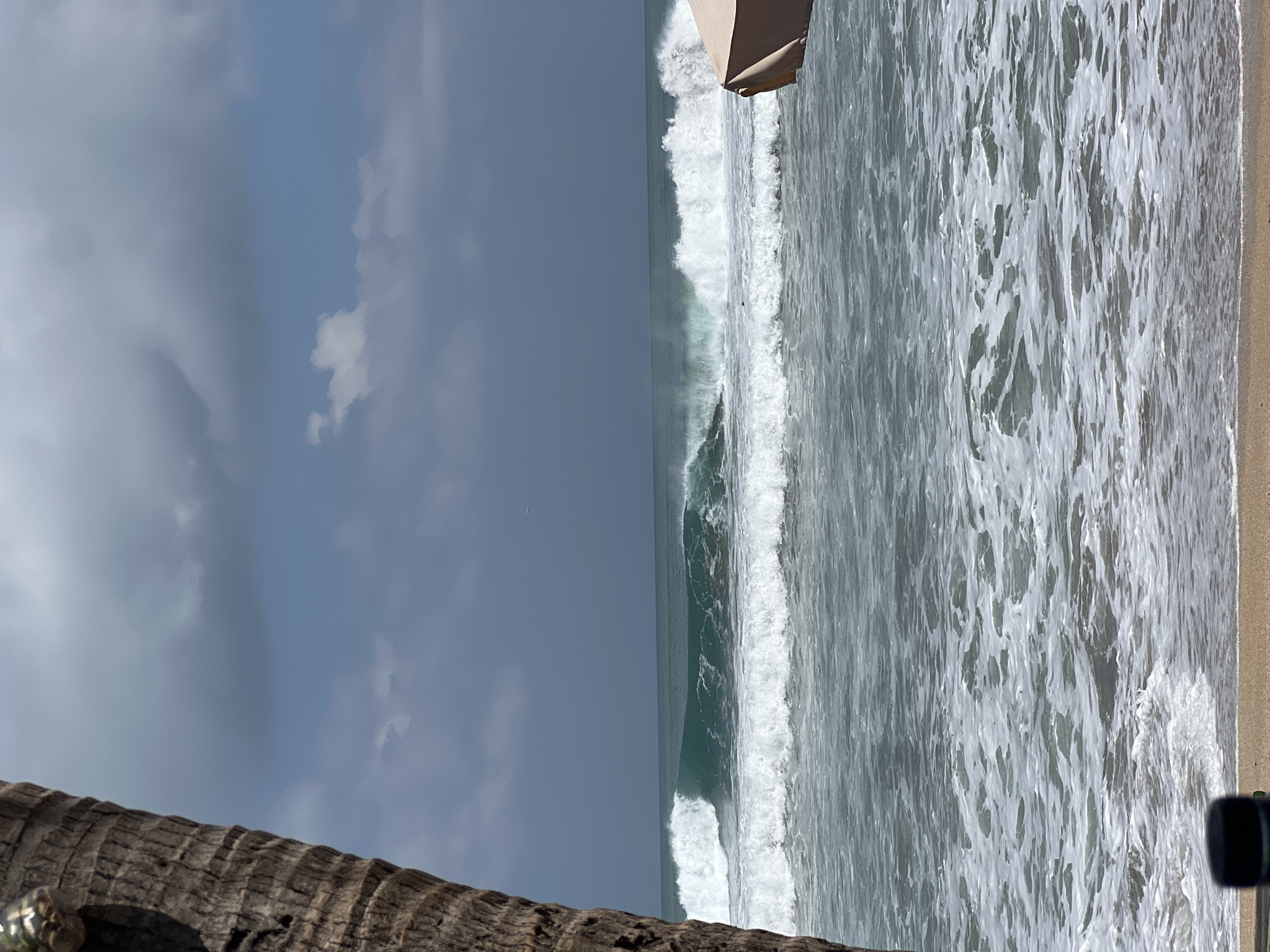 A close-up of a palm tree trunk occupies the left foreground of the image. The trunk is textured and rough, with visible horizontal lines and a dark brown color. Beyond the trunk, the scene opens to a view of the ocean and the sky. Several waves are breaking in the mid-ground. The waves are a deep teal color as they crest and then transition to white foam as they crash onto the shore. In the background, the sky is a mix of dark gray clouds and patches of lighter blue, with some small white clouds scattered throughout. The horizon line is clearly visible, separating the sea from the sky. On the right side of the image, a portion of a light tan beach umbrella is visible. The foreground also shows a sandy beach with some darker objects at the very bottom. The overall environment suggests a tropical beach setting in Kuta, Indonesia. There is no indication of any specific actions or interactions taking place beyond the natural movement of the waves.