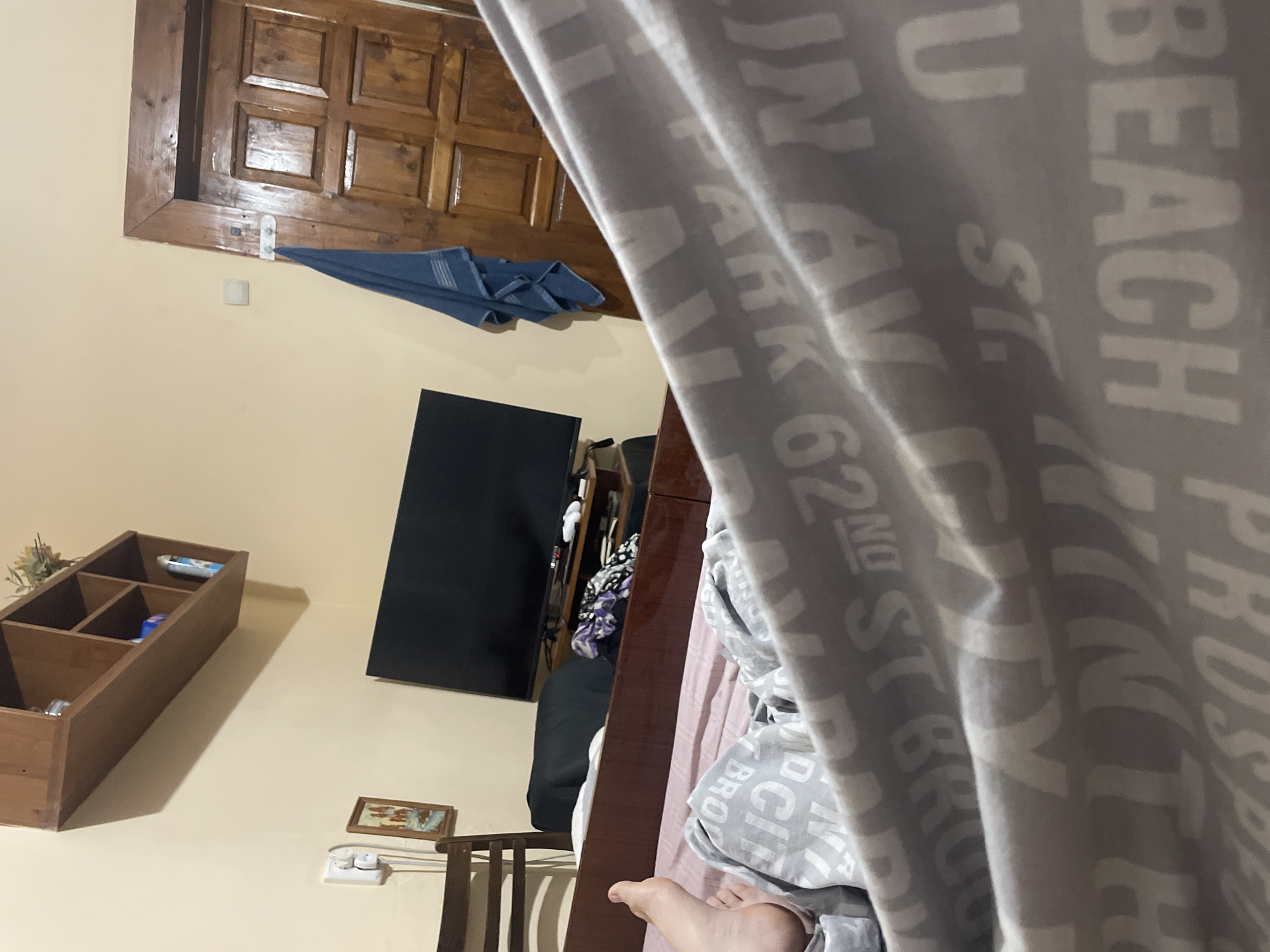 The image depicts a dimly lit bedroom interior, likely in an apartment or house. In the foreground, a person's left foot is visible, resting on a bed covered with a blanket featuring a repeating pattern of text that appears to be street names and numbers. The bed frame is made of dark wood.
In the mid-ground, a dark wooden chair is positioned against a light yellow wall. A small framed picture hangs on the wall above the chair. Further back, a black television is mounted on a dark wooden media console. A pile of folded clothes in various patterns lies on the console. A blue towel is hanging from a hook on a wooden door in the background, which has a multi-paneled design. Above the television, a wooden shelf unit is mounted on the wall, containing a few items, including what appears to be a spray can and some decorative elements. The overall impression is of a lived-in, somewhat cluttered space. There is no indication of the specific location within the city of Chernivtsi, Ukraine, beyond the context provided by the prompt.