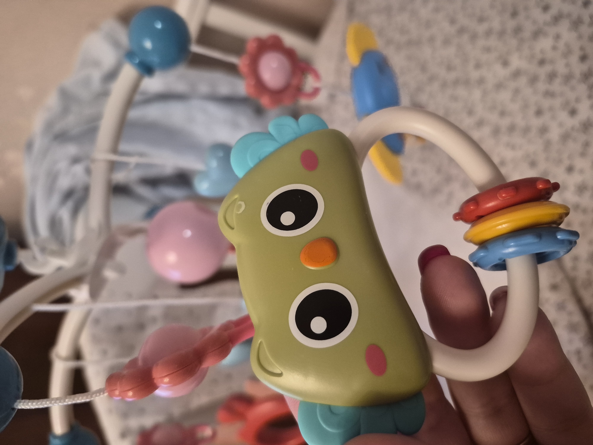 A close-up shot depicts a colorful baby mobile toy. The central figure is a green, stylized animal head with large, black-and-white eyes and a small orange nose. It has turquoise accents on its head and chin. The toy is suspended from a white curved arm with red, yellow, and blue rings. A hand with painted fingernails is holding the toy. In the blurred background, other elements of the mobile can be seen, including blue spheres, pink flowers, and other colorful shapes. The backdrop is a neutral, textured wall or surface. There is no indication of Tobolsk, Russia.