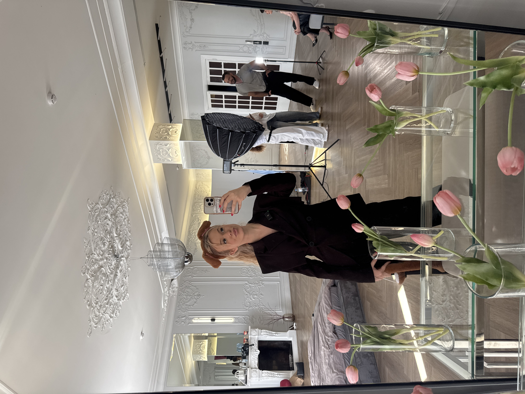 A young woman with blonde hair is taking a selfie in a large mirror. She is wearing a dark blazer and brown dog ear headwear. In the foreground of the mirror, there are several glass vases filled with pink tulips. In the background, reflected in the mirror, there is a studio setting. A man is posing near a softbox light, and two other people are standing behind him. Further in the background, there are French doors, a white fireplace, and a bed. The room has ornate white wall moldings and a decorative ceiling medallion. The flooring is light wood. The overall impression is that of a photoshoot in an elegantly decorated studio space.