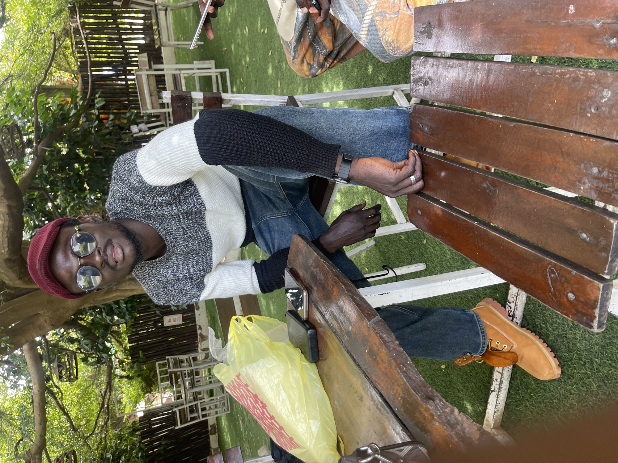 The image depicts an outdoor seating area, likely a restaurant or cafe, in Jos, Nigeria. A young Black man, wearing a red cap, sunglasses, and a grey and white striped sweater with black sleeves, sits at a wooden table. He is wearing blue jeans and tan work boots. On the table, there is a yellow plastic bag, two cell phones, and a wallet. Another person's legs, wearing patterned trousers, are visible on the right side of the frame, also seated at a table. The environment features lush green grass, trees providing shade, and outdoor furniture, suggesting a relaxed atmosphere. The lighting indicates it is daytime, with sunlight filtering through the leaves.