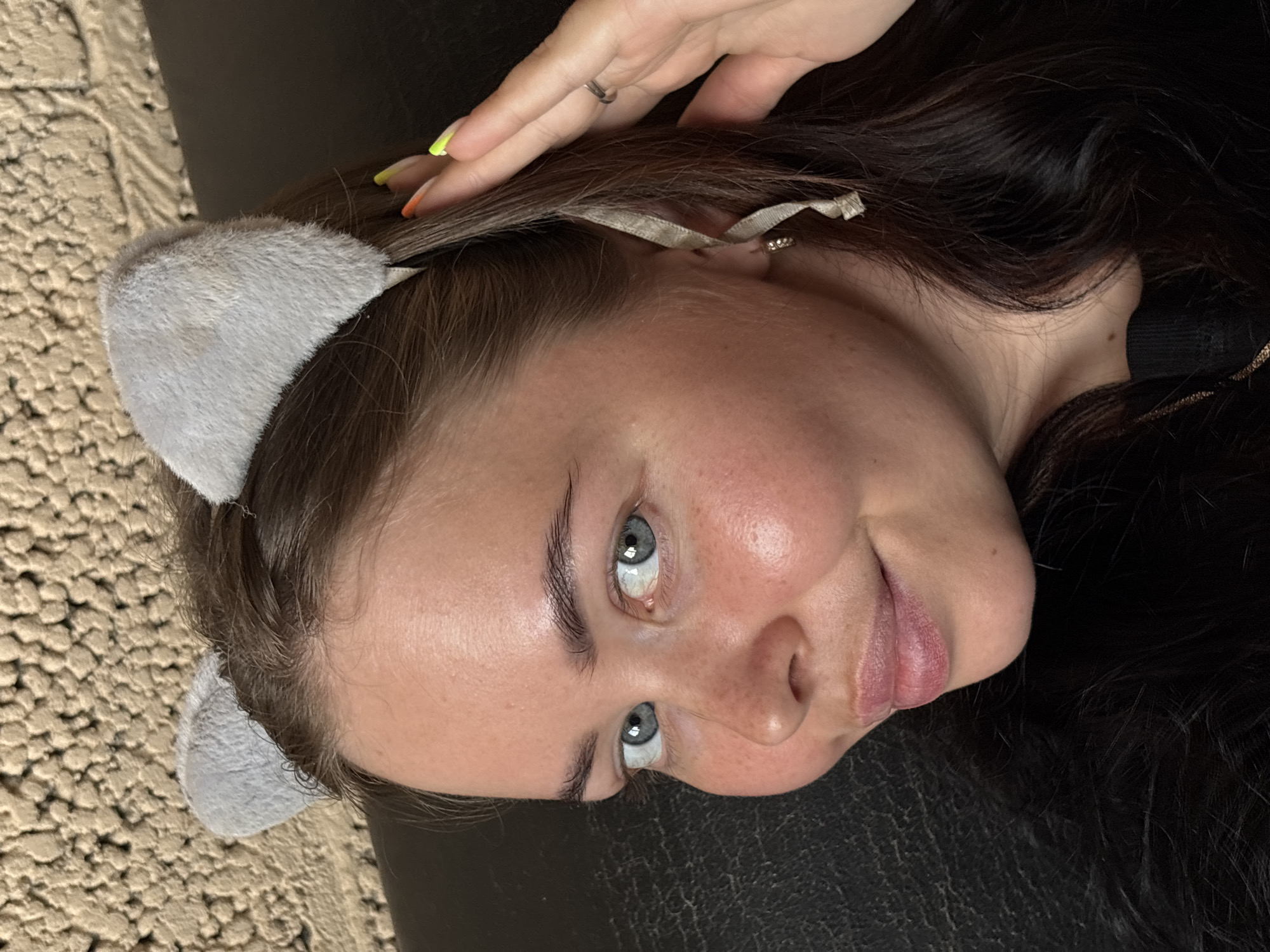 A woman lies with her head on a black leather surface. She wears gray cat ears. Her dark hair falls around her face, and a hand with painted nails rests on her forehead. She looks up at the camera. The background on the left is a textured beige surface. No information indicates a location of Русский Вожой, Russia.
