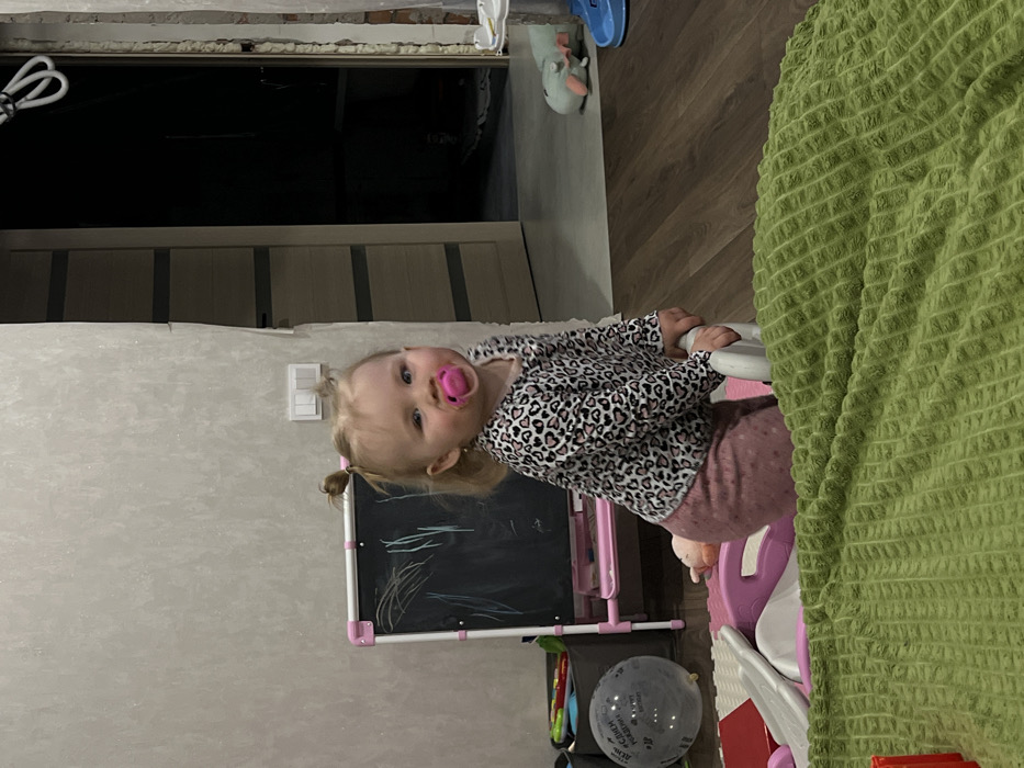 A young child with light-colored hair in two pigtails stands in an indoor residential setting in Leninakan, Russia. The child, wearing a long-sleeved black and pink heart-patterned top and pink pants, holds a pink pacifier in their mouth. They grasp a white, curved portion of a child's toy, possibly a walker or small play equipment. A small earring is visible in the child's left ear.
The immediate foreground features a dark green, textured blanket or mat. To the child's left, part of a pink and white plastic play structure rests on white interlocking foam floor tiles. Further left, a grey storage bin contains various colorful objects, and a grey balloon with "С ДНЁМ РОЖДЕНИЯ" (Happy Birthday) text is visible. Behind these items, a child's easel with a pink frame and a blackboard surface displays white chalk scribbles.
The room has light-colored, textured walls. A white light switch with two toggles is mounted on the wall to the right of the easel. A wooden door with horizontal stripe details is partially open, revealing a dark, unlit room beyond. To the right of the door, an exposed wall section indicates renovation, showing brickwork and white expanding foam around a doorway. On the dark wood laminate floor to the right, a blue plastic toy and a light-colored plush animal toy are present. The floor throughout the visible area is dark wood laminate.