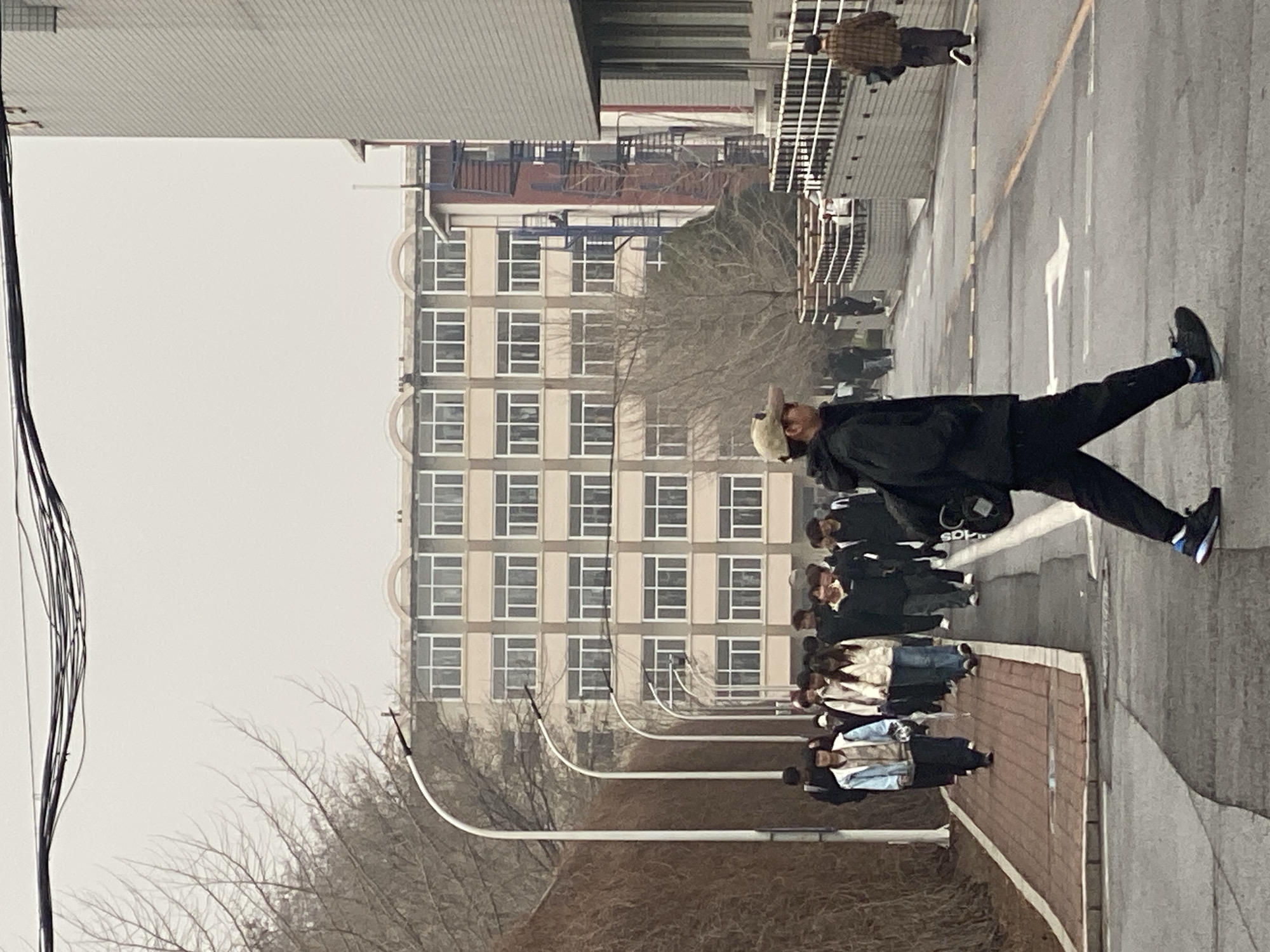 The image shows a group of people walking along a street in Zhongtun, China. The street is lined with trees and streetlights. In the background, there is a large building with many windows. The sky is overcast. A person is walking to the right side of the camera.