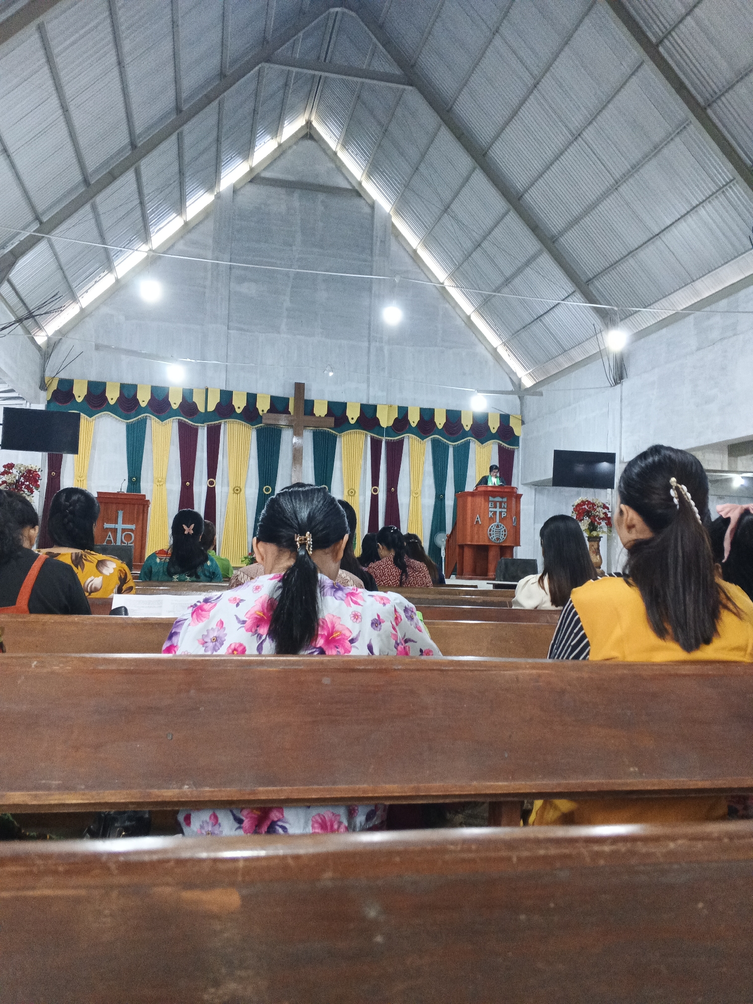 The image depicts the interior of a church in Limau Manis, Indonesia. The perspective is from the back of the congregation, looking towards the altar. The ceiling is constructed with corrugated metal panels supported by exposed beams, creating a distinctive A-frame structure. Natural light streams in through gaps in the roofline, and artificial lights are also visible.
In the foreground, wooden pews are occupied by several individuals, mostly women, seen from the back. They are dressed in casual attire. One woman in the immediate foreground wears a white top with a floral pattern of pink and purple flowers. To her right, another woman is wearing a yellow top.
At the front of the church, there is a pulpit with a cross on it, and a banner with colorful fabric drapes. A person, likely a preacher or speaker, stands behind the pulpit. There is a large television screen mounted on the wall behind the pulpit. A vase of red flowers sits on a stand to the left of the pulpit. The walls appear to be concrete. The overall atmosphere suggests a religious service or gathering.