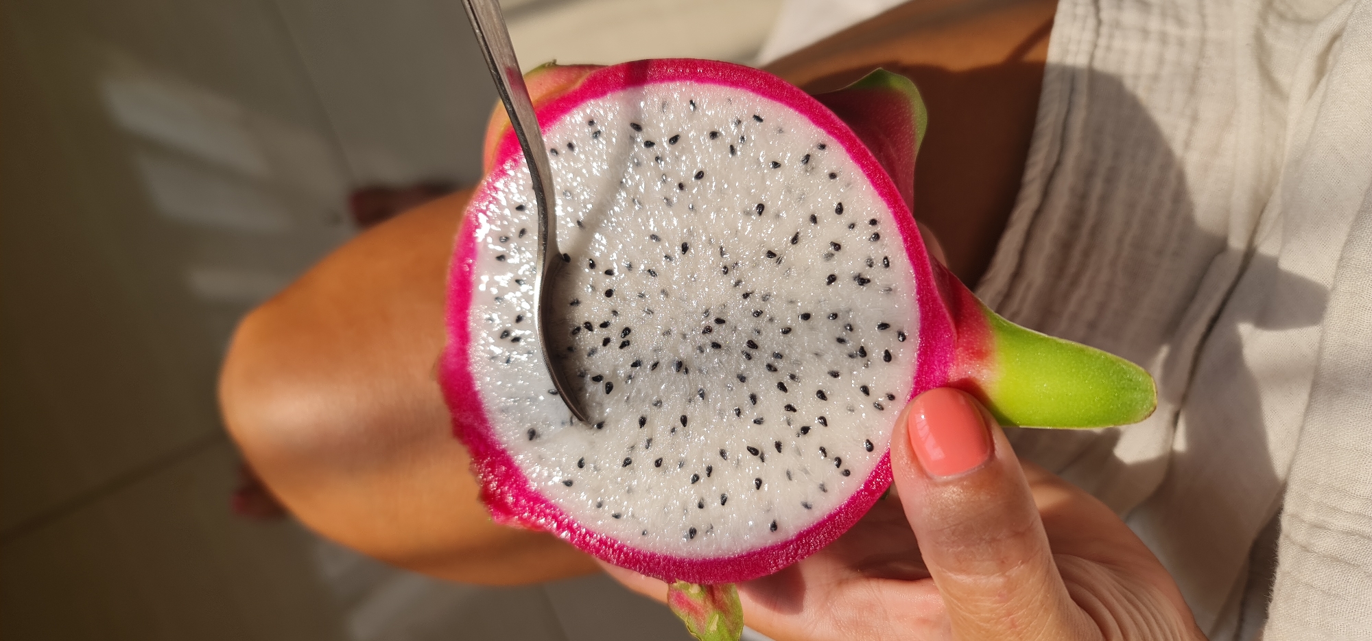 A close-up shows a halved white dragon fruit held by a hand with coral-colored nail polish. A spoon is inserted into the white pulp with black seeds. The fruit's pink outer rind and green stem are visible. The person's skin tone is tan. Part of a light-colored fabric garment is visible. The location is unspecified but inferred to be in Đà Nẵng, Vietnam.