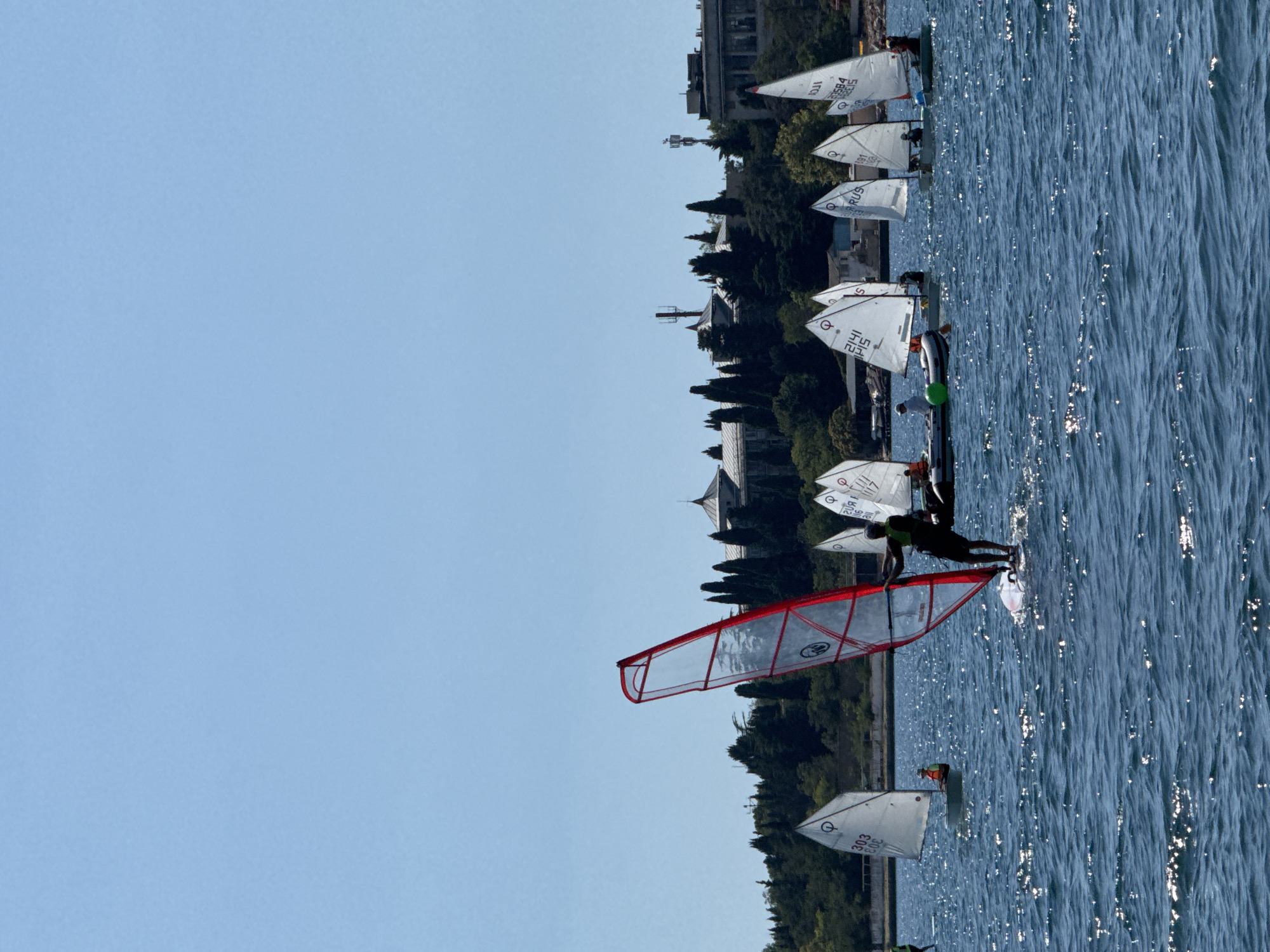 On a sunny day, a fleet of small sailboats and a windsurfer are on the water. The windsurfer, in the foreground, is on a white board with a red and white sail. Several sailboats, with white triangular sails, are scattered across the water. In the background, a tree-covered hill overlooks the water, with several buildings and communication towers visible on top. The buildings appear to be a complex of historical or institutional architecture. The water is choppy and reflects the sunlight. This scene is likely taking place in the vicinity of Sevastopol, Ukraine, given the architectural style of the buildings and the maritime activity.