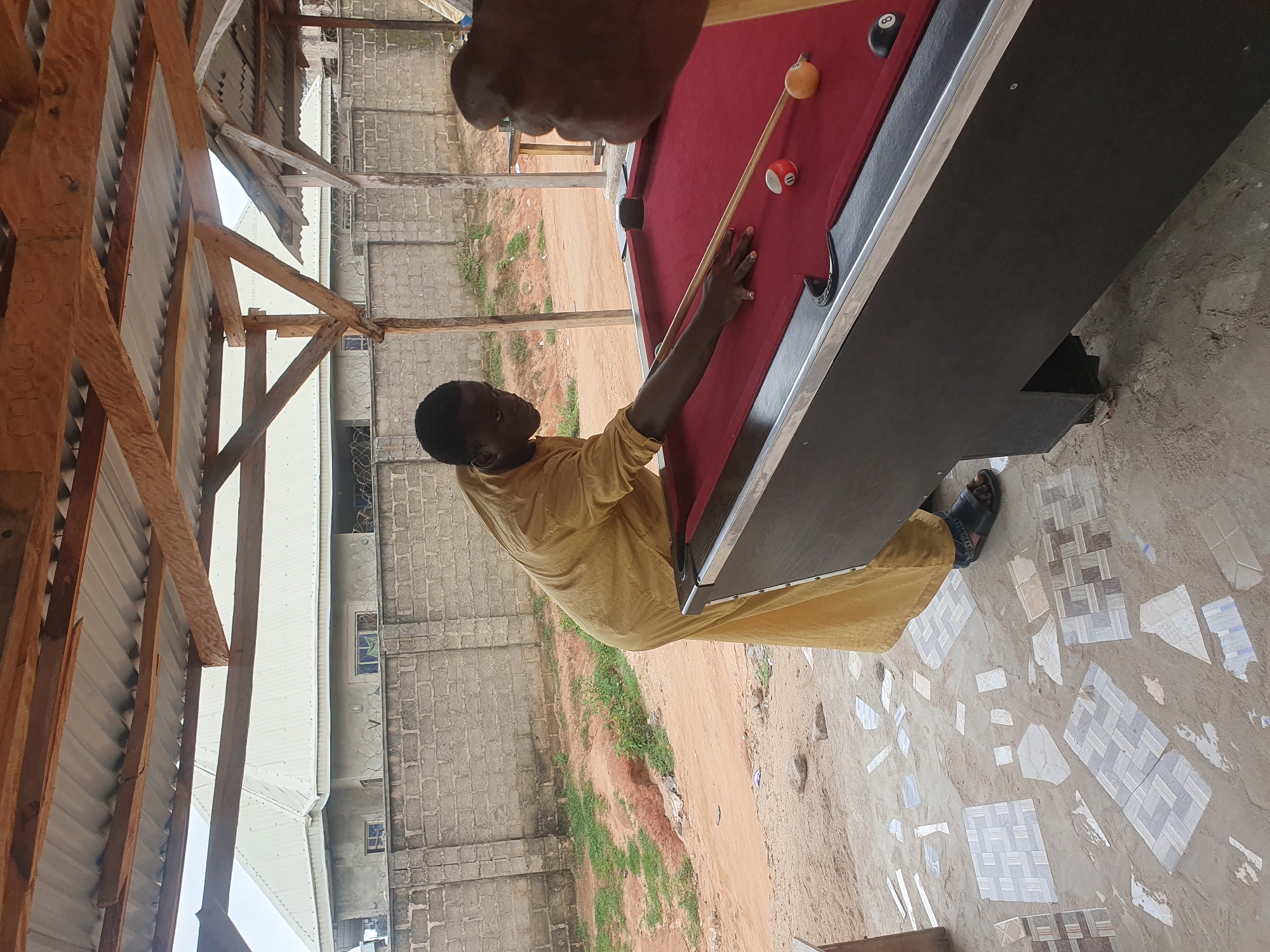 In Lokoja, Nigeria, a young Black male, wearing a yellow robe and sandals, plays pool under an open-air structure with a corrugated metal roof supported by wooden beams. He is positioned to strike a cue ball with a pool cue, aiming at another ball on a red felt billiard table. A second person's hand and arm, holding a pool cue, is visible on the right side of the frame, suggesting they are also participating in the game. The ground surrounding the pool table is a mixture of dirt and scattered mosaic tiles, indicating an unfinished or rustic outdoor area. In the background, a brick wall and a building with a white metal roof are visible. The overall scene depicts a casual game of pool in an outdoor setting in Lokoja.