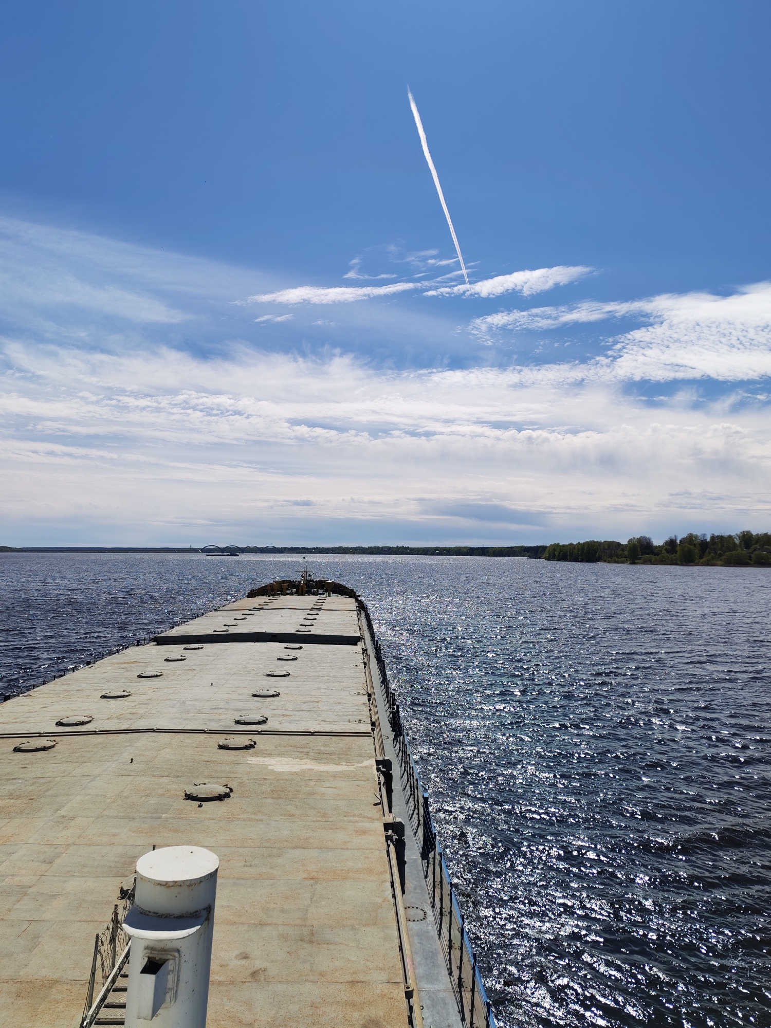 The image shows a view from a boat or dock, looking out over a body of water. A flat, concrete surface with hatch covers stretches out into the water. To the right and left of this surface is the water, sparkling in the sunlight. In the distance is a shoreline with trees and possibly a bridge. The sky is blue with scattered clouds and a contrail from an airplane. The location is identified as Glebovo, Russia.