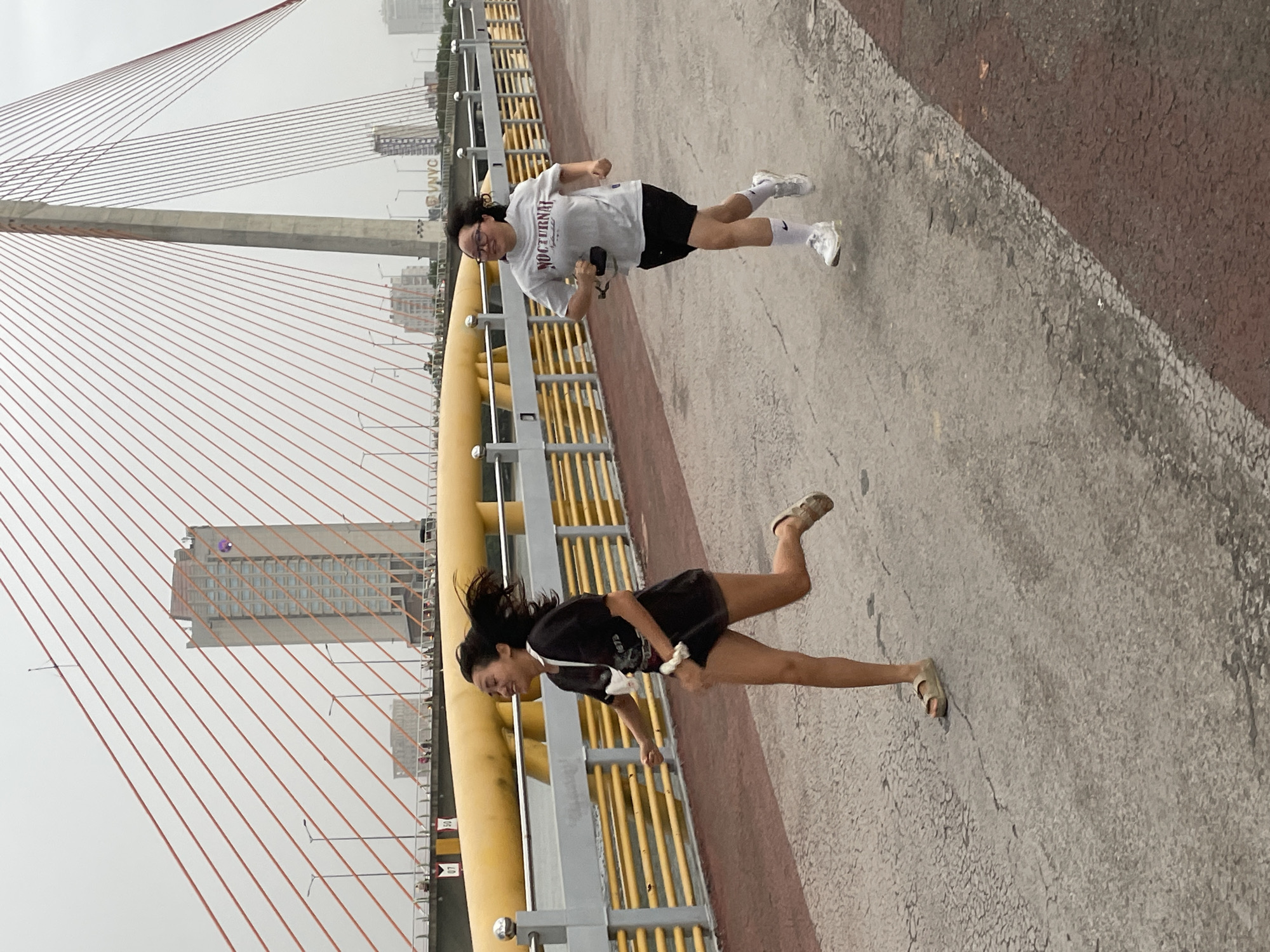 This image depicts two young women running on a cable-stayed bridge, likely in Đà Nẵng, Vietnam, given the architectural style and surrounding urban landscape. The bridge features prominent yellow support structures and numerous red cables stretching across the frame. In the background, modern buildings and a hazy, overcast sky suggest a daytime scene, possibly in the morning or afternoon.
The woman in the foreground is wearing a black t-shirt and shorts, with her hair flowing behind her as she runs. She appears to be smiling and engaged in the activity. The woman behind her is wearing a light-colored t-shirt with "NOCTURNAL" printed on it and black shorts. She is also running, holding a phone in one hand, and looking towards the camera with a slight smile. Both are wearing athletic shoes.
The activity taking place is clearly exercise, specifically running, on a pedestrian walkway of the bridge. The ground surface is a mix of concrete and asphalt. The overall mood appears active and pleasant, with the subjects enjoying their run outdoors. The overcast weather may indicate a humid climate, common in this region.