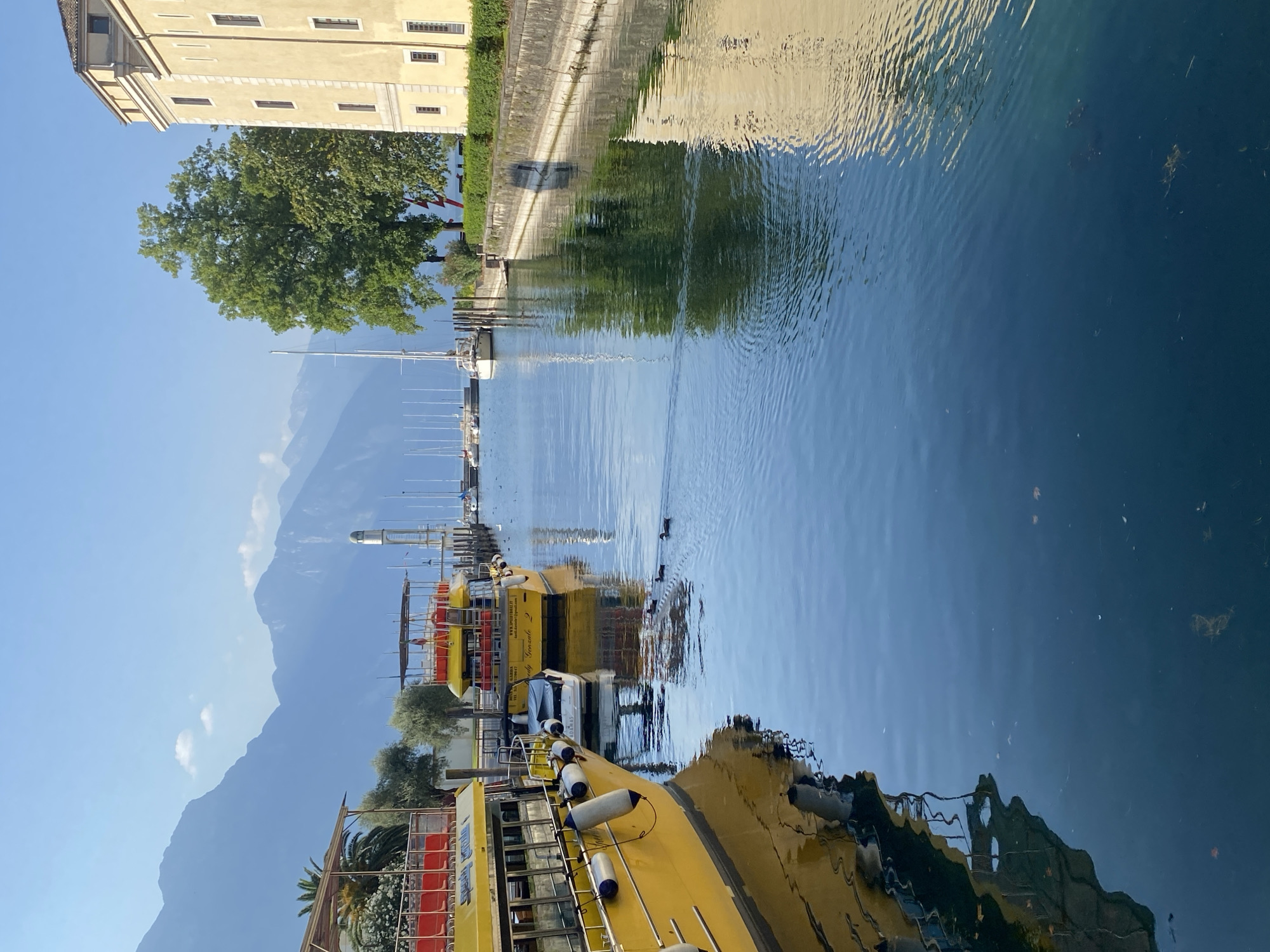 The image depicts a canal in Riva del Garda, Italy, with a view towards the mountains. On the left, a yellow boat with red seating is docked. Further down the canal, another yellow boat is also docked, and a smaller white boat is moored between the two. The water is calm and reflects the boats and the surrounding buildings and trees. In the distance, sailboats are visible, and beyond them, a lighthouse stands tall. The background features a range of mountains under a clear blue sky. On the right side of the canal, a pale yellow building with a tiled roof is situated along the embankment, which is lined with green bushes. Several ducks are swimming in the water. The scene appears to be peaceful and picturesque, capturing the charm of the lakeside town.