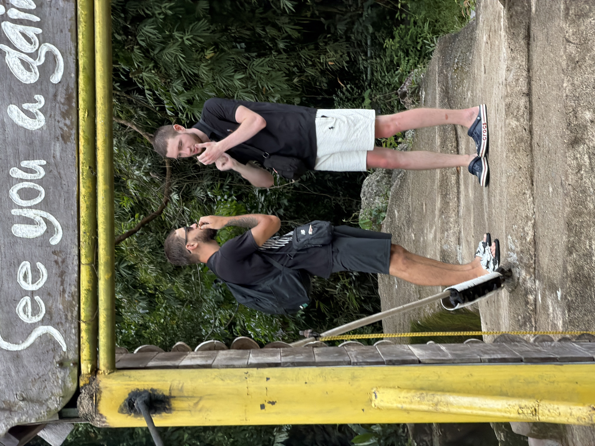 Two young adult males are standing on a set of concrete stairs. The male on the left is wearing a black t-shirt, black shorts, a black backpack, and black and white patterned flip-flops. He has sunglasses perched on his head and is holding his hand to his mouth. The male on the right is wearing a black polo shirt, white shorts, and dark blue Crocs. He has a small black crossbody bag slung over his shoulder and is holding his hands up near his chin. Both individuals have short hair. They are standing under a wooden sign that reads "See you again" in white lettering. A yellow metal structure with a chain hanging from it is to the left of the frame. The background consists of dense green foliage, suggesting an outdoor, possibly tropical, environment. The scene appears to be in Khanh Vinh, Vietnam, based on the information provided.