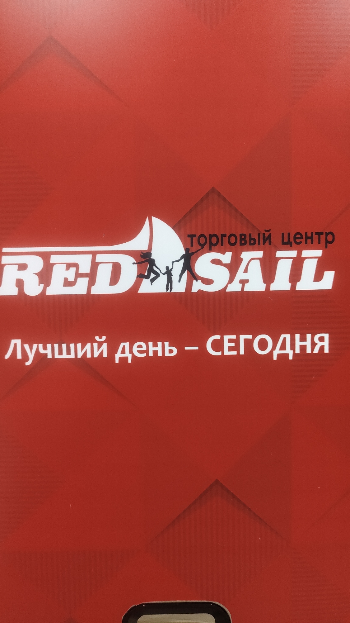The image shows a red banner with white text and graphics. The text reads "RED SAIL" in large white letters, with stylized silhouettes of people running and jumping above the letters "D" and "S". Above "RED SAIL" is smaller text in Russian that says "ТОРГОВЫЙ ЦЕНТР", meaning "shopping center". Below "RED SAIL" is more Russian text that says "Лучший день - СЕГОДНЯ", meaning "The best day - TODAY". The banner appears to be an advertisement or logo. Without further information or context, the specific location in Krasnoyarsk, Russia, cannot be determined from the image alone.