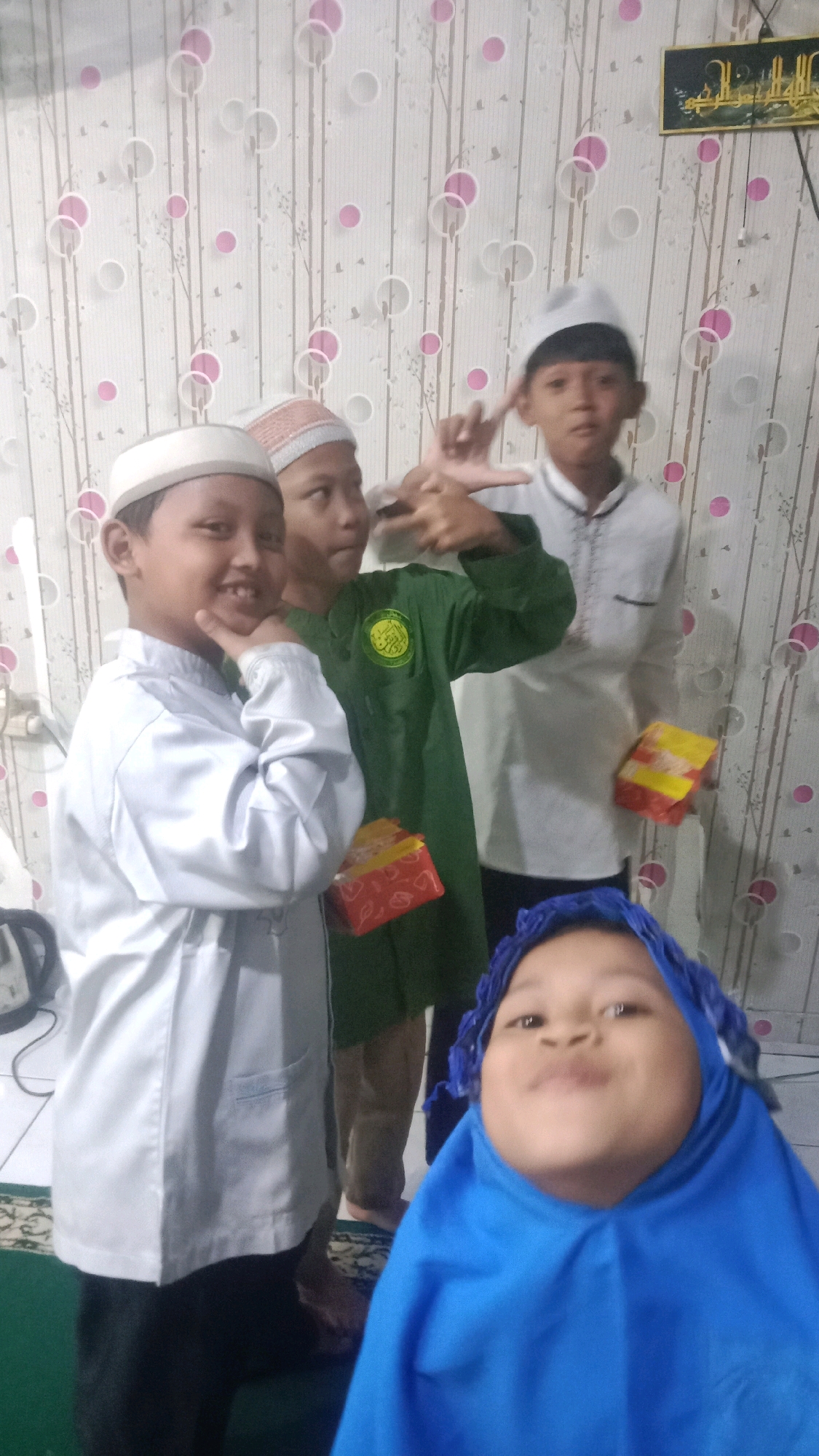 In the city of Sumberjaya, Indonesia, a group of children is pictured in a room with floral-patterned wallpaper. A young boy in the foreground, wearing a white cap and a silver outfit, smiles as he touches his chin with his hand. Behind him, another boy in a green shirt holds a gift box. To the right, a boy in a white shirt with traditional embroidery also holds a gift box. In the very front, a child wearing a blue hijab looks directly at the camera with a playful expression. The environment appears to be indoors, possibly a home, with a patterned rug visible on the floor.