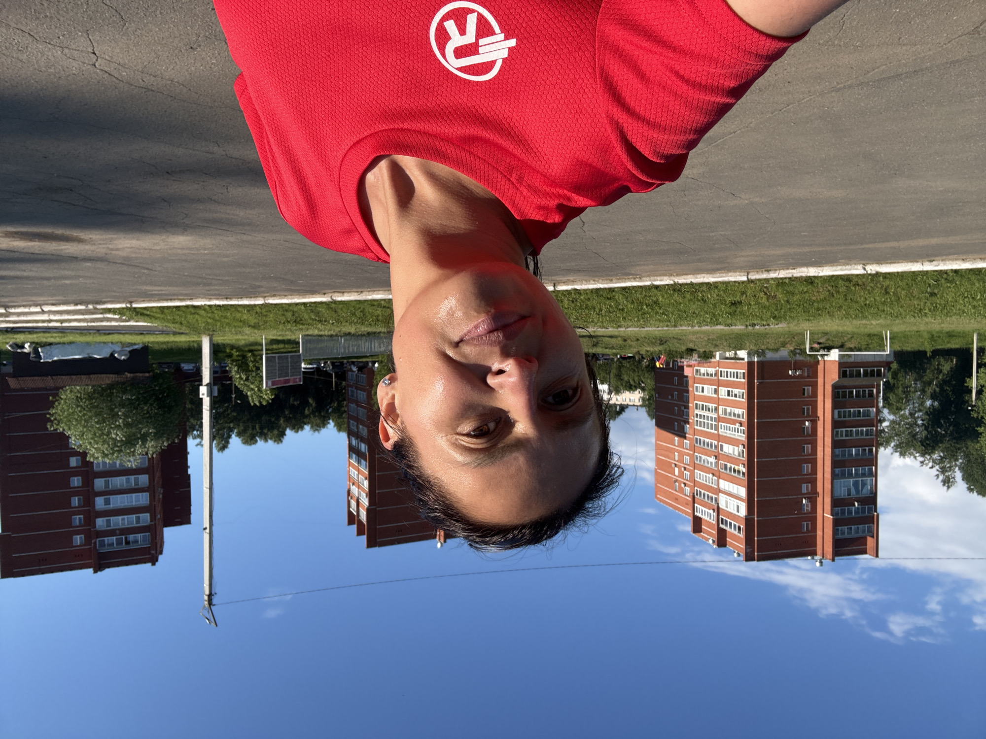 The image depicts a woman taking a selfie outdoors in Chaikovsky, Russia. She is shown from the chest up, looking directly at the camera with a neutral to slightly content expression. Her face has a subtle sheen, suggesting recent physical activity. She wears a bright red athletic t-shirt made of textured fabric. On the left side of her chest, a white logo is visible, featuring a stylized letter "R" combined with a horizontal bar and an arrowhead.
Behind her, an urban residential scene unfolds under a clear blue sky adorned with a few white clouds. Two multi-story brick apartment buildings, each with multiple white-framed windows, stand prominently in the mid-ground on either side of the frame. A lush green tree partially obscures the building on the left. A utility pole with overhead wires extends vertically in the center. In the foreground, beneath the woman, there's a paved asphalt road or path, bordered by a strip of green grass. The bright sunlight indicates it is daytime, likely morning or afternoon.