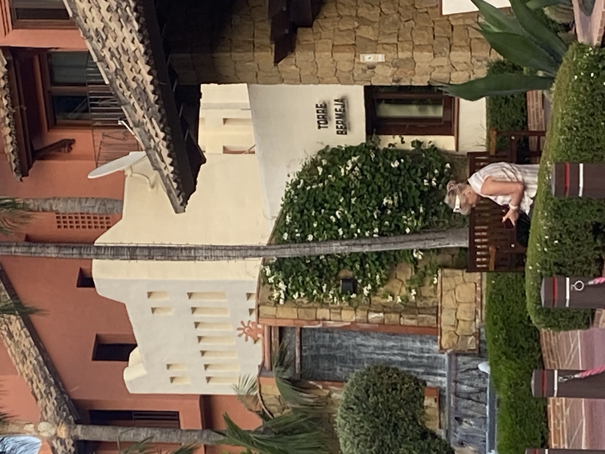 The image depicts the exterior of the Torre Bermeja development in Estepona, Spain. A woman is seated on a wooden bench in the foreground, looking at a red object in her hands. She is wearing a striped sleeveless shirt and a white headband. Behind her, a lush green vine with white flowers climbs a wall, adjacent to a cascading waterfall feature. The architecture is characteristic of the region, with terracotta tile roofs and warm-toned stucco walls. The scene is set in an outdoor area, likely a courtyard or garden, with manicured hedges and palm trees visible.
