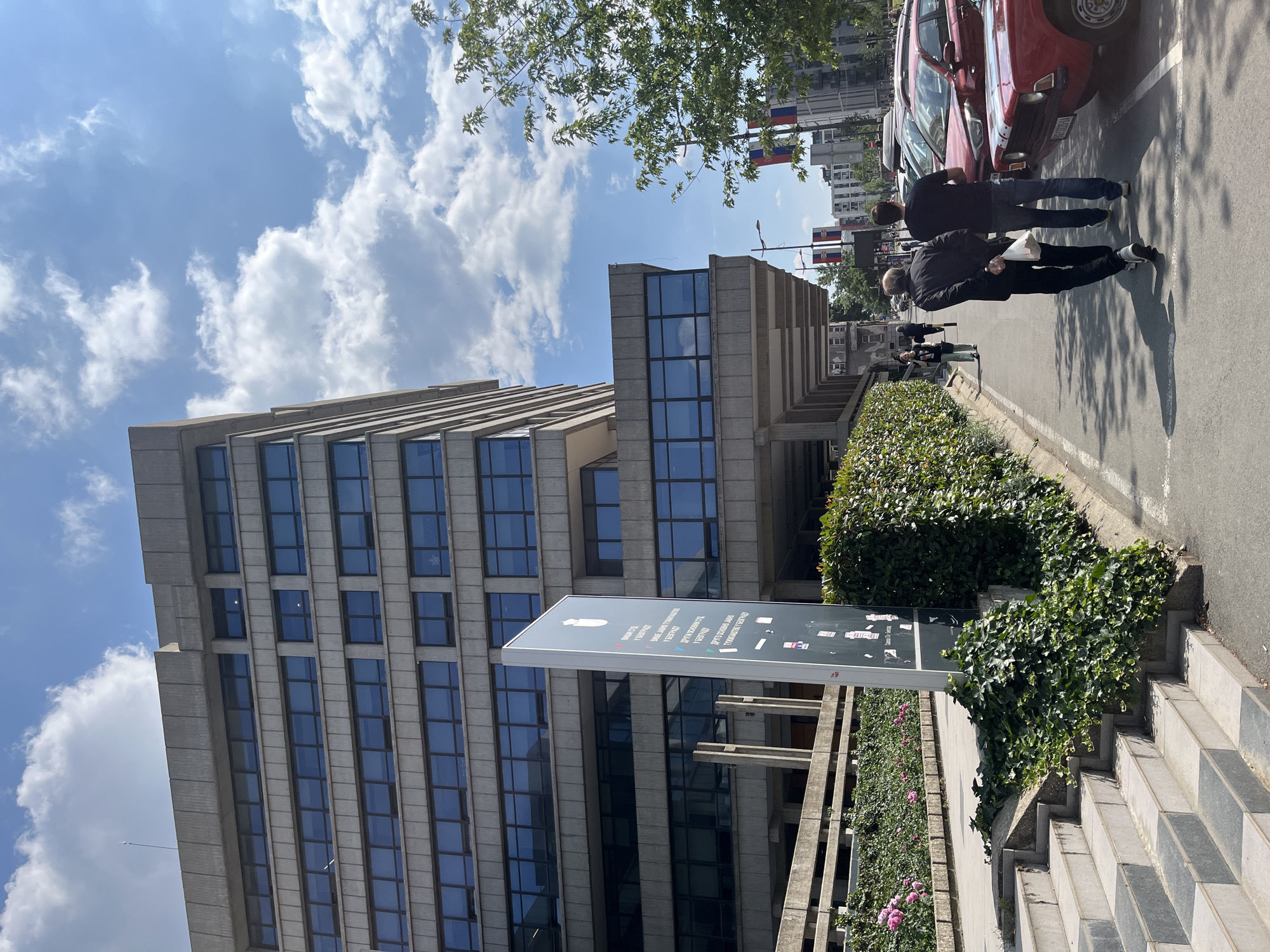 An urban street scene unfolds on a bright day, featuring a prominent, tall concrete building with numerous dark, rectangular windows, suggestive of a Brutalist architectural style. The building dominates the left side of the frame, its structure characterized by strong horizontal and vertical lines.
In the foreground, a large, vertical grey sign stands, bearing text in what appears to be Serbian Cyrillic and Latin script, including words like "SLOBODA" (Freedom), "ZAKONA" (Law), and "BEZBEDNOST" (Security), indicating a public or governmental institution. Lush green hedges and ivy line the pavement and the base of the building, with some pink flowers visible among the greenery. Stone steps lead up from the sidewalk on the far right.
On the asphalt street to the right, two men are walking away from the camera. The man in the foreground wears a dark jacket and trousers, carrying a white bag. Behind him, another man in a dark shirt and blue jeans walks beside a parked red car, which appears to be an older model, possibly a Yugo, a common sight in the region. Other vehicles are parked further down the street.
The sky above is bright blue with scattered white, fluffy clouds. Overhead, green tree branches and leaves are visible, adding a natural element to the urban setting. Shadows cast on the ground suggest it is daytime, likely midday or early afternoon. In the background, visible through gaps in the trees and buildings, are flags with red, blue, and white stripes, consistent with the Serbian flag, reinforcing the Belgrade context. The overall atmosphere is one of a typical bustling day in a city.
