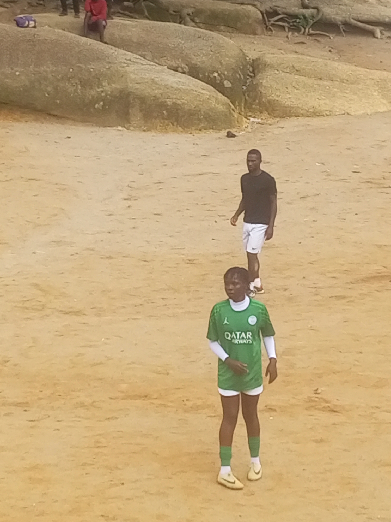 Two individuals are on a sandy, outdoor terrain with large rock formations in the background. In the foreground, a person wearing a green soccer jersey with "QATAR AIRWAYS" visible, white shorts, white socks, and yellow cleats stands facing the camera. They have dark skin and short, dark hair. Behind and to the left of this person, another individual is visible, wearing a black t-shirt and white shorts, also with dark skin and short dark hair, facing to the left. The terrain appears to be sandy or dusty, with some small debris scattered. The background shows large, weathered rocks and tree roots. It is unclear if this location is specifically in Jos, Nigeria, as there are no identifiable landmarks.