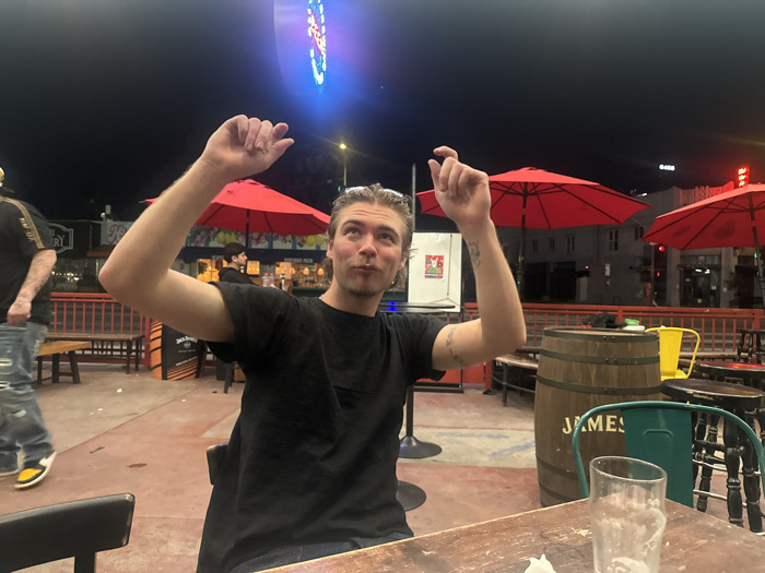 This image depicts an outdoor patio at Jumbo's Clown Room, located at 5173 Hollywood Boulevard, Los Angeles, California, United States of America, during nighttime hours.
The central subject is a young adult male with light-colored, medium-length hair, wearing a plain black t-shirt and blue jeans. He is seated at a rustic wooden table, with both arms raised above his head, hands slightly open, and fingers bent. His facial expression indicates a reaction, with his mouth slightly open and eyes directed upwards toward the sky. A tattoo is visible on his left forearm. An empty, clear drinking glass with residue rests on the table in front of him.
To the left, a second individual is partially visible, walking away from the camera, wearing a dark shirt with gold accents, ripped light blue jeans, and yellow sneakers. A third individual is seated or standing in the mid-ground, under a large red patio umbrella, wearing a dark hoodie. Multiple other indistinct figures are present in the background.
The patio features a red-toned concrete floor, bordered by red metal railings. Several open red patio umbrellas are distributed across the area. Seating options include wooden benches, yellow metal chairs, green metal chairs, and dark bar stools. A wooden barrel with "JAMES" written vertically on its side is positioned next to a green chair. An orange sign displaying "JACK DANIELS" branding is visible on the left. In the background, part of a sign reading "BREWERY" is discernible. The dark sky is illuminated by a prominent, multi-colored neon sign, glowing blue and red, featuring a distinctive stylized crown logo, confirming the establishment as Jumbo's Clown Room. Other buildings are visible in the distance, including one faintly displaying the number "5450," and additional red neon signage. The scene is artificially lit, characteristic of an active nightlife venue.